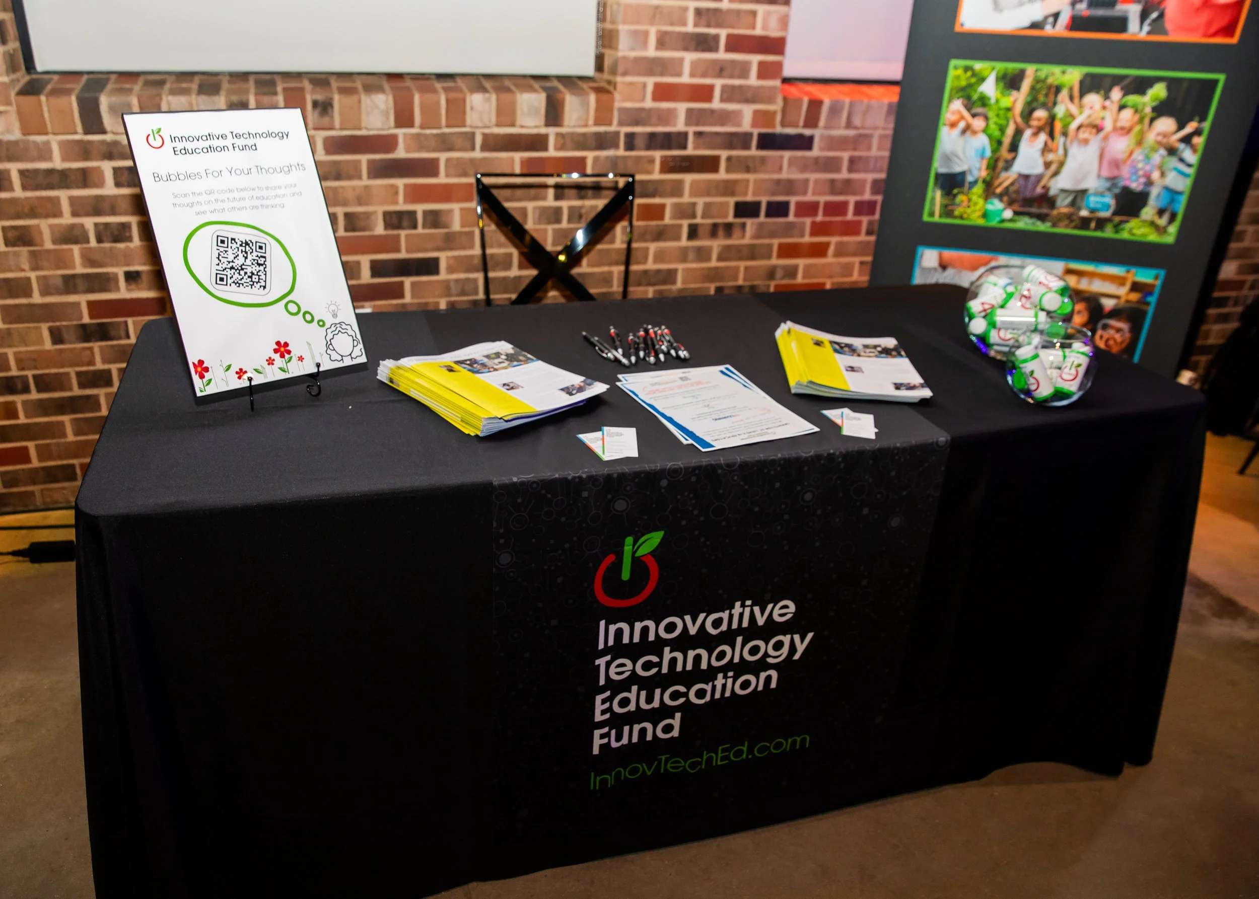 A table with promotional materials for Innovative Technology Education Fund, including brochures, pens, a bowl of small bottles, and a sign with a QR code, set against a brick wall.