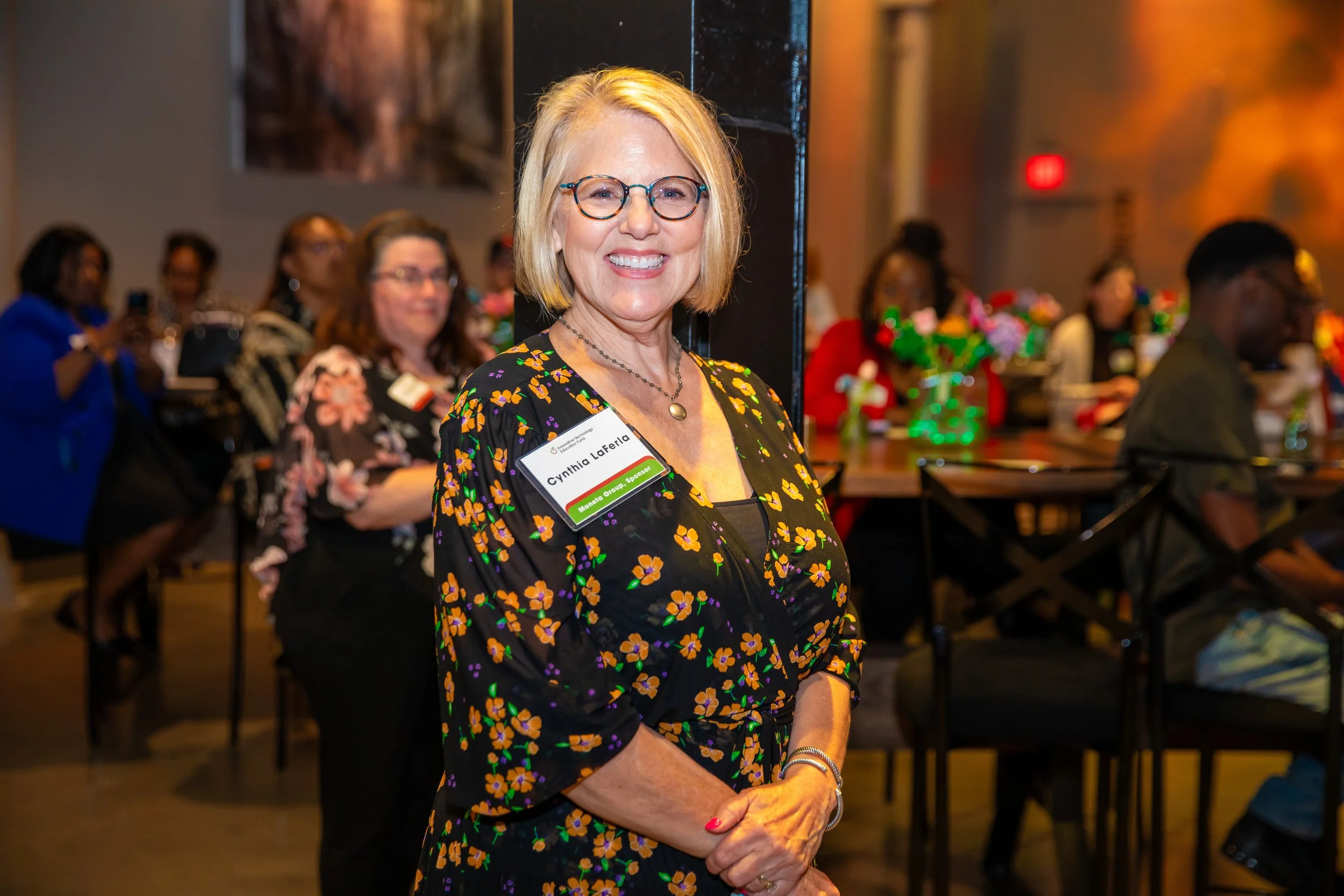 A woman wearing glasses and a floral dress smiling at a social gathering. She has a name tag reading 'Cynthia LaFera' and 'Sponsor'. There are people in the background, some taking photos, and the setting appears to be indoors, possibly at a conferen