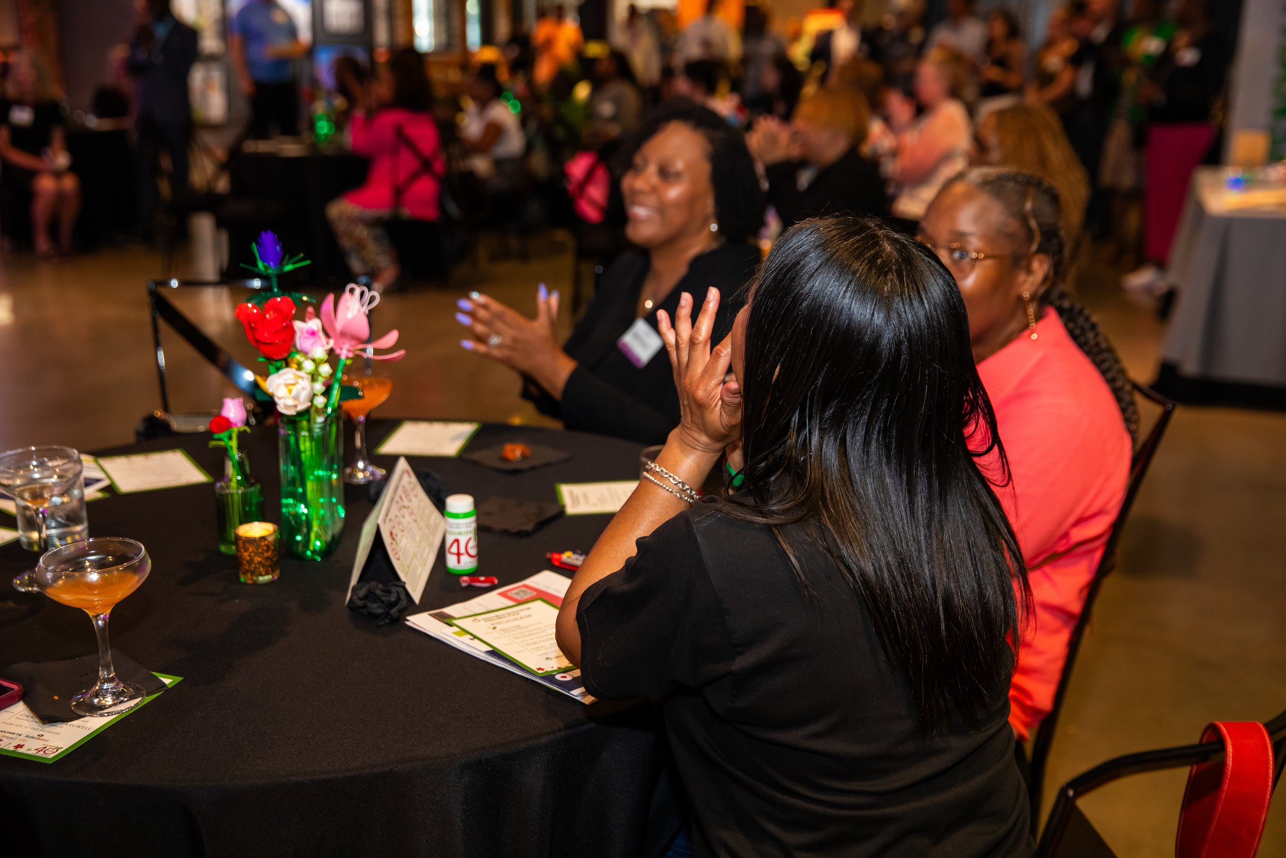 People sitting at a round table in a lively indoor setting, smiling and engaging in conversation, with colorful decorations, drinks, and informational pamphlets on the table.
