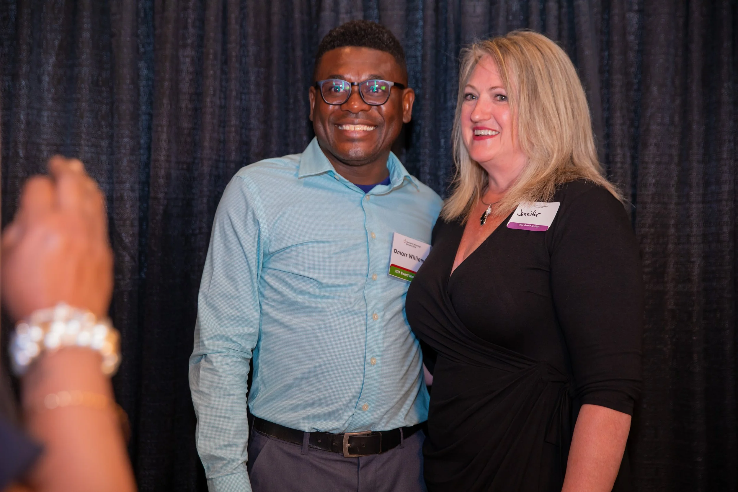 A man and a woman standing together, smiling, at an indoor event with a dark curtain backdrop. The man is wearing glasses and a light blue shirt, and the woman has blond hair and is wearing a black blazer with a name tag.
