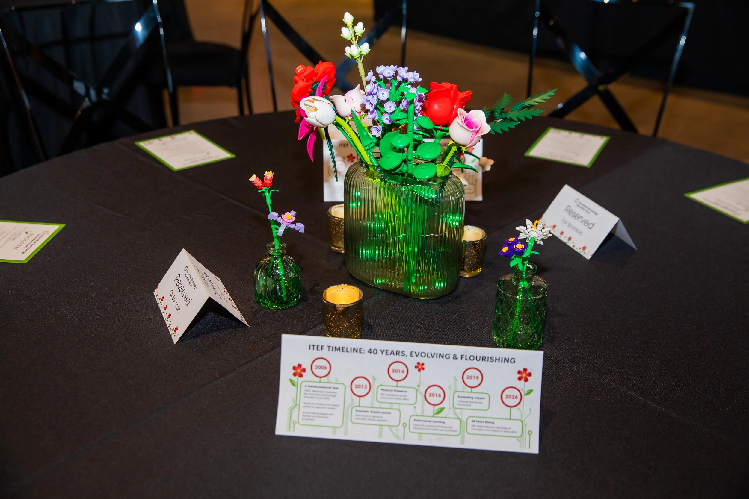 A floral arrangement of plastic LEGO flowers in a green glass vase on a table, surrounded by smaller vases with similar flowers and lit candles. There are informational signs and reserved signs on the table.