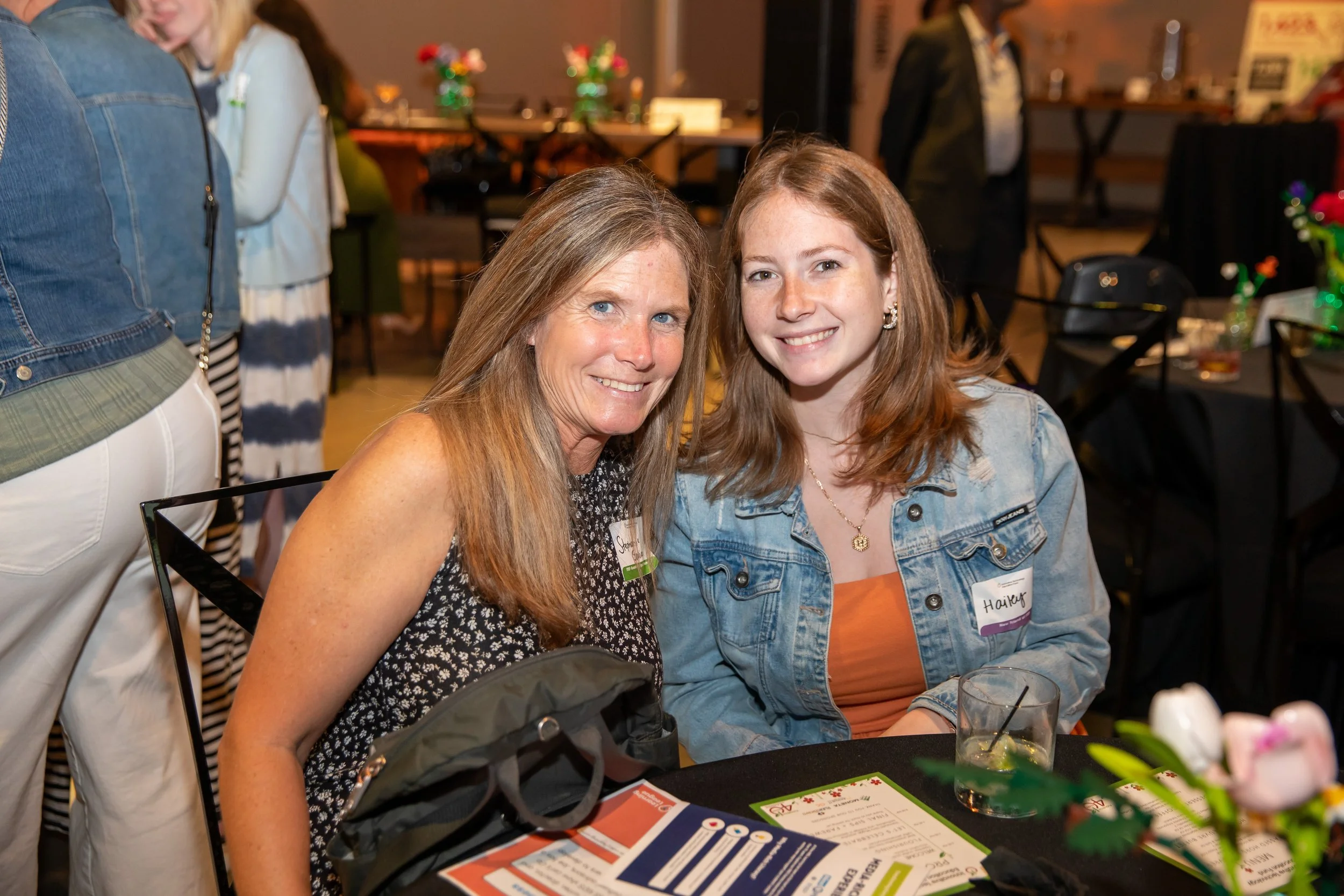 A woman and a young girl sitting at a table at an event, smiling at the camera. The table has informational pamphlets and a drink. Other people and tables are visible in the background.