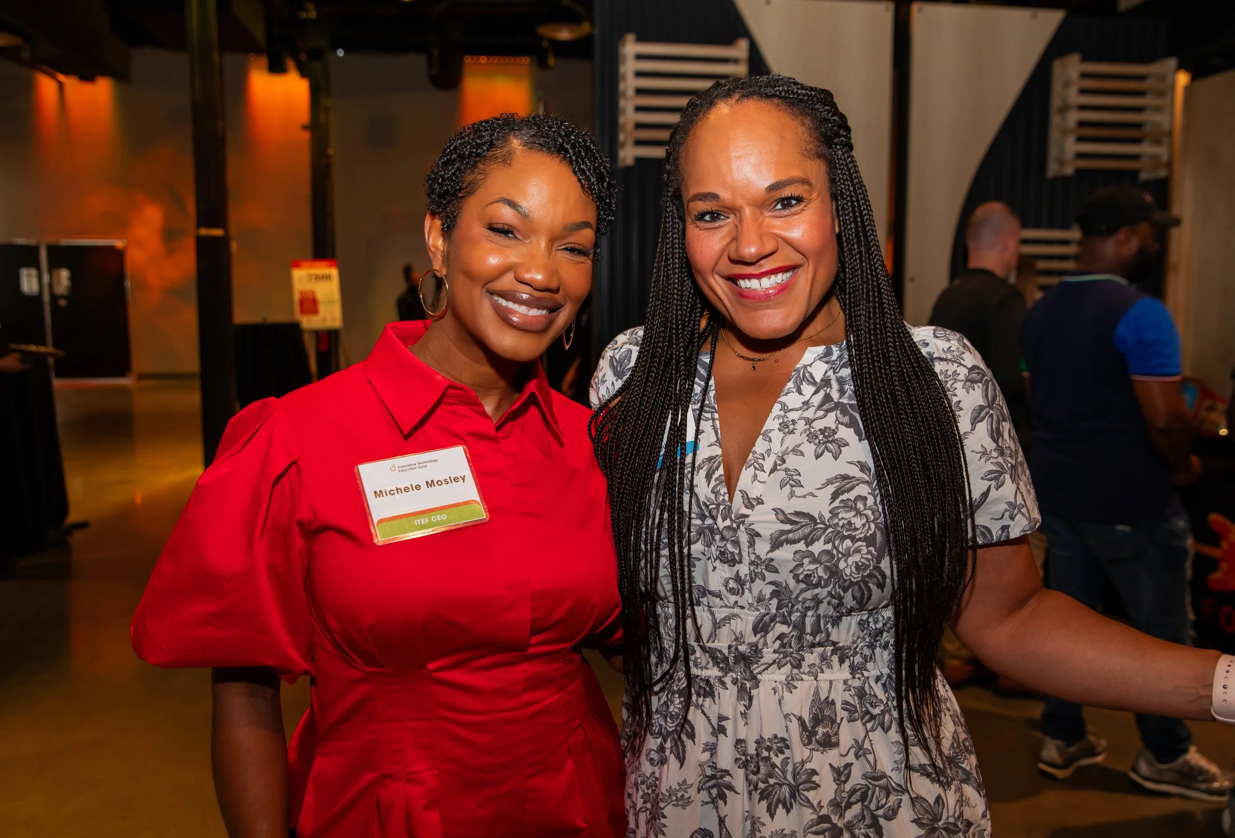 Two smiling women standing closely together at an indoor event, with people and background decor visible behind them.