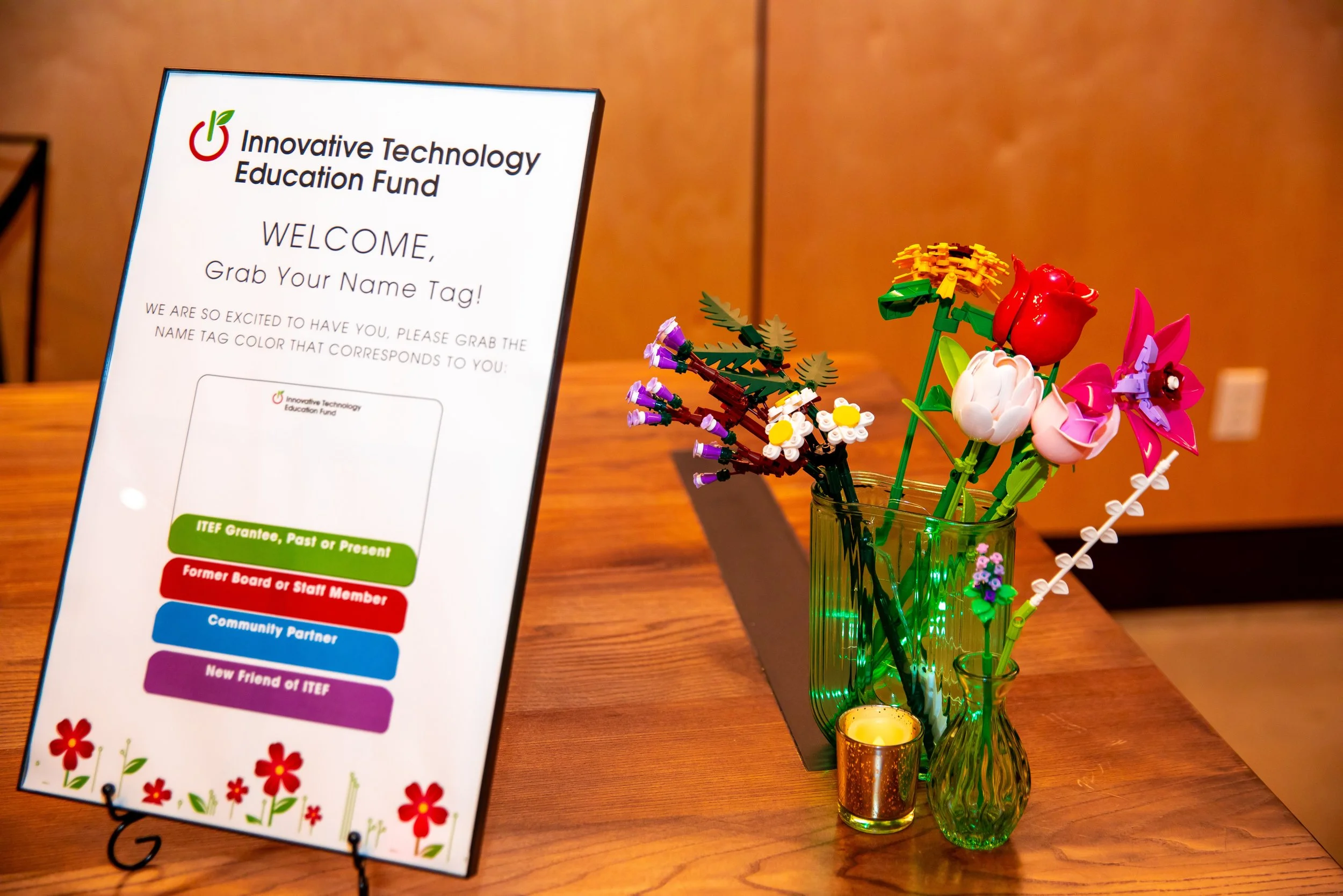 A sign for the Innovative Technology Education Fund with instructions for name tags. Next to the sign is a glass vase holding colorful artificial flowers, a small lit candle holder, and a purple flower decoration, arranged on a wooden table.