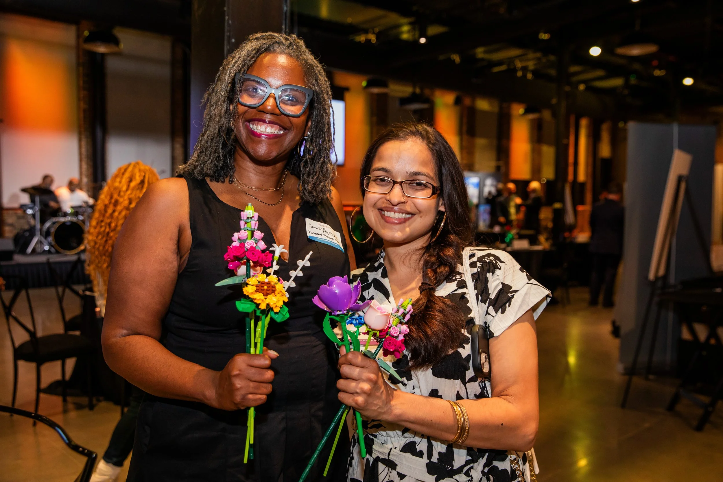 Two women smiling and holding colorful flower-themed Lego bouquets at an indoor event. One woman has curly gray hair, glasses, and a black sleeveless top; the other has long dark hair, glasses, and a black-and-white patterned dress.