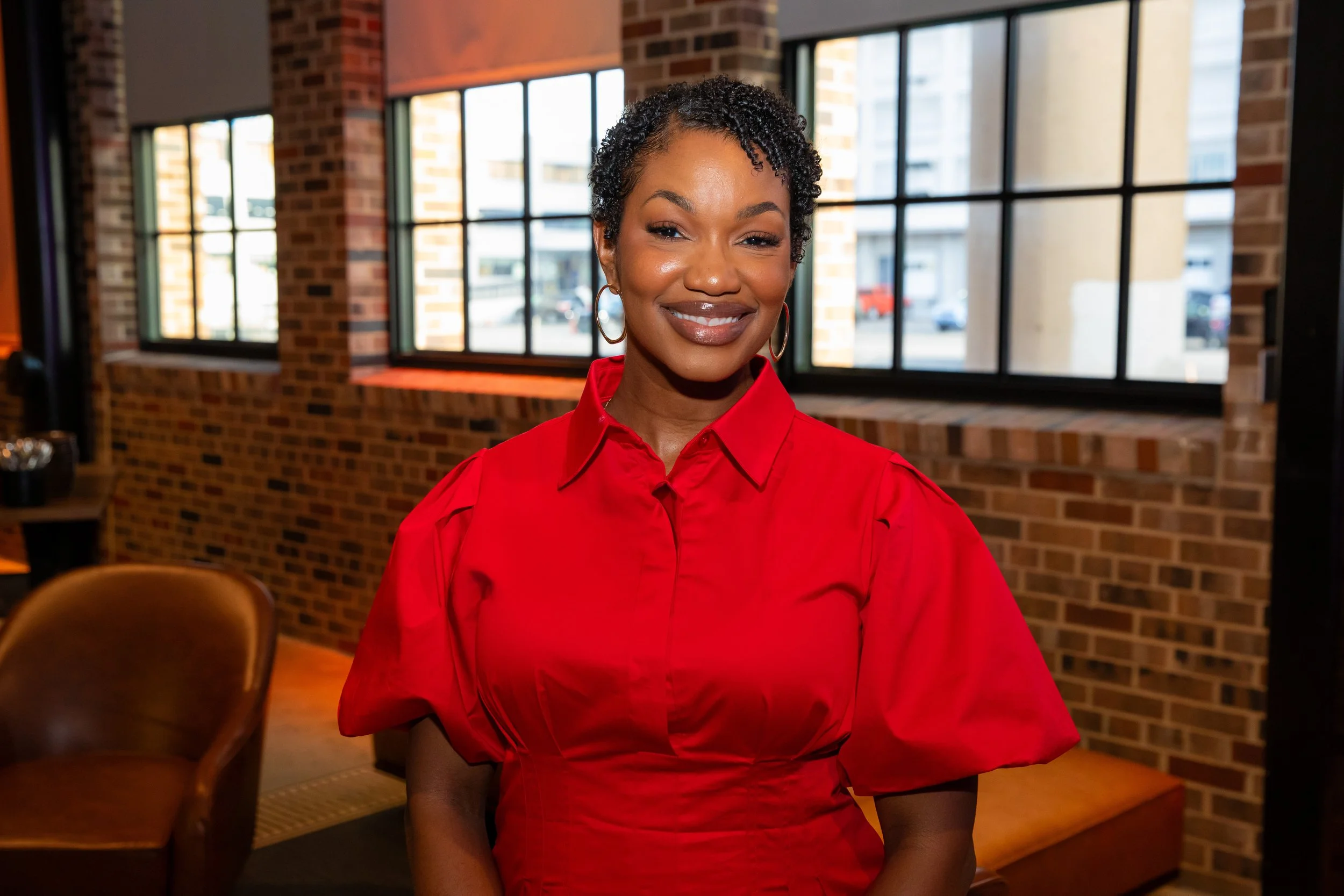 A woman with short curly hair and hoop earrings wearing a red blouse, smiling in a room with brick walls and large windows.