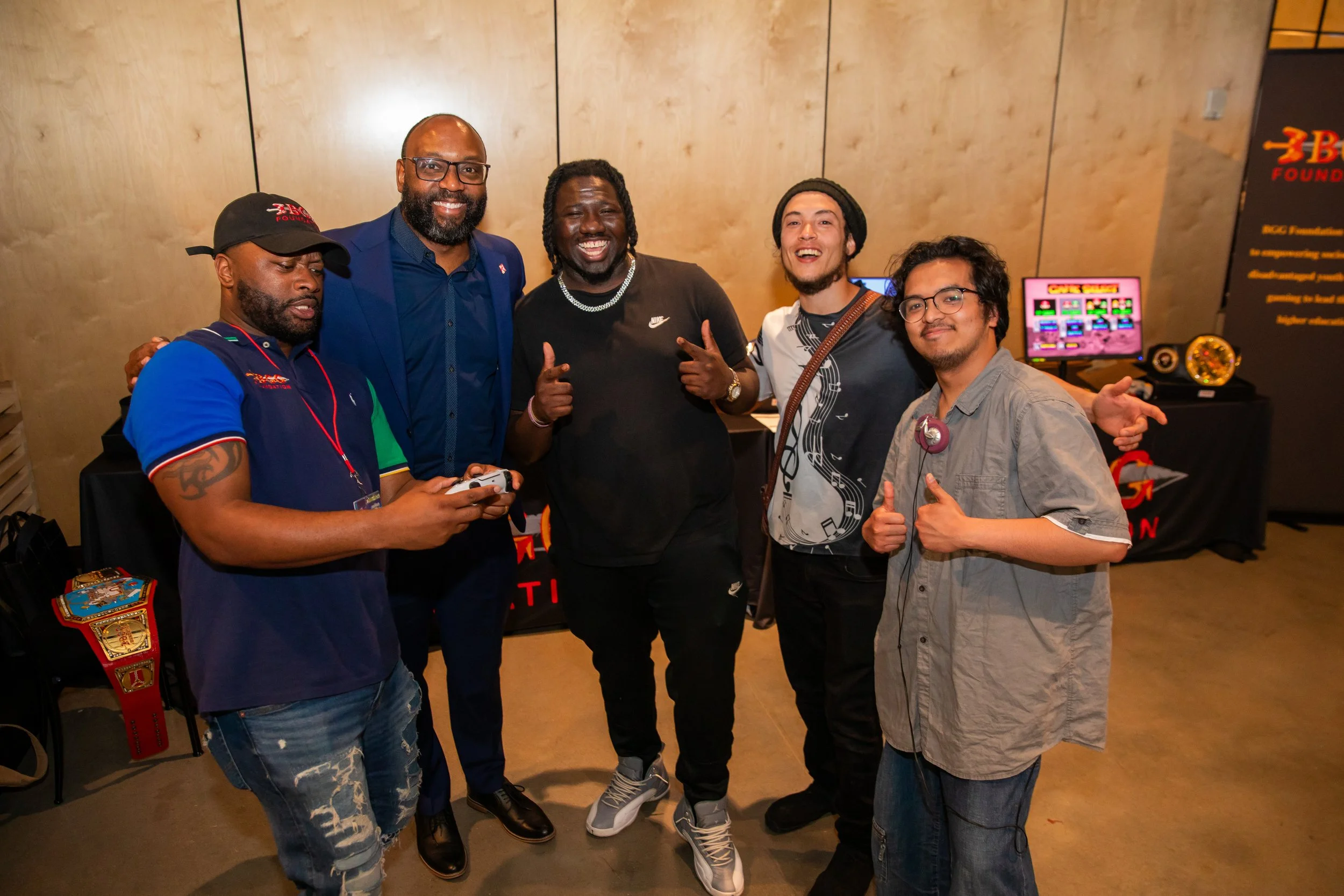 Group of five men standing together indoors, smiling for the photo. Four are wearing casual clothing, and one in the middle is dressed in a blue suit and glasses. Background features a beige wall and a gaming machine.