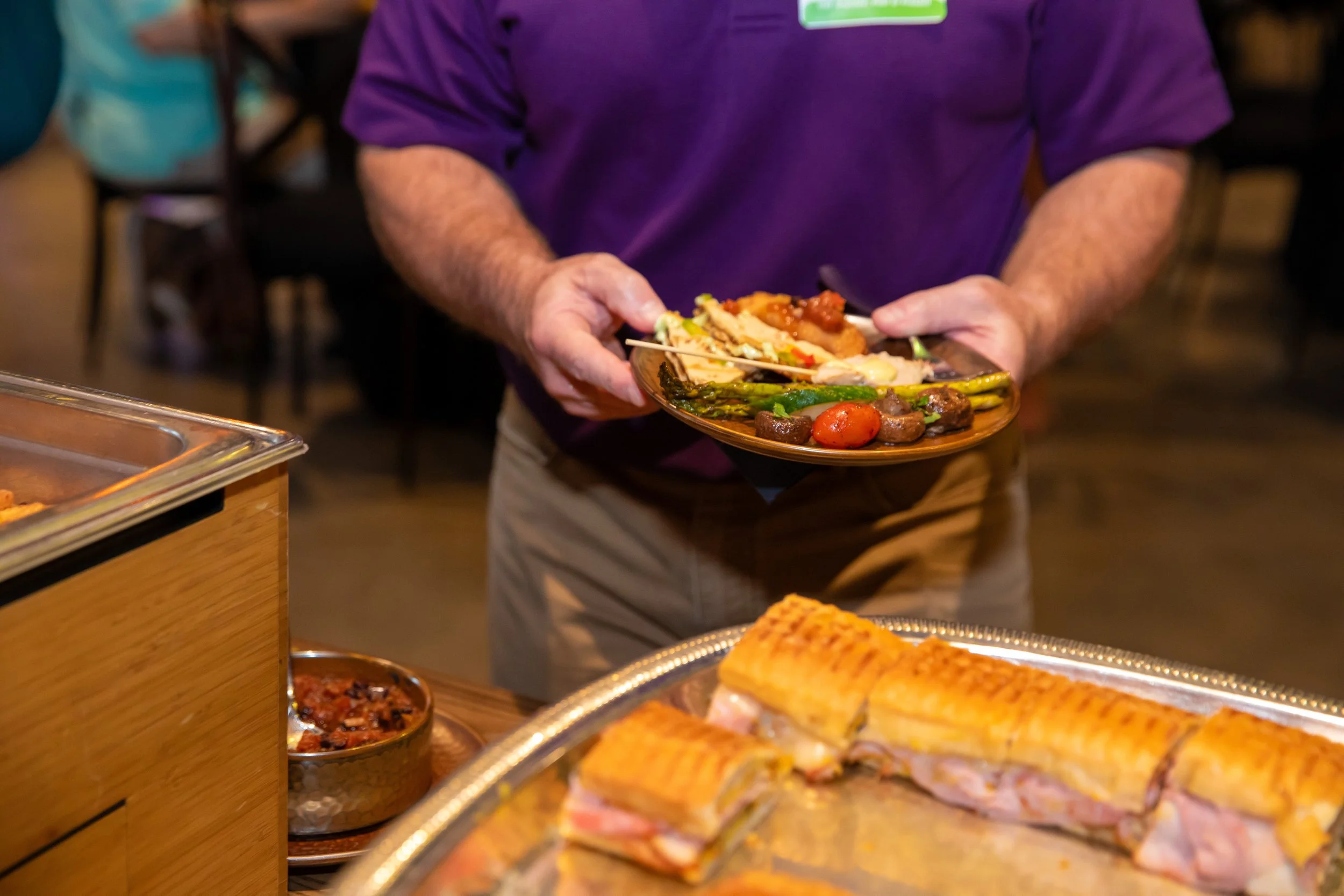 A person in a purple shirt holding a plate of assorted food, including vegetables and grilled items, with various dishes and food items on a buffet table in front.