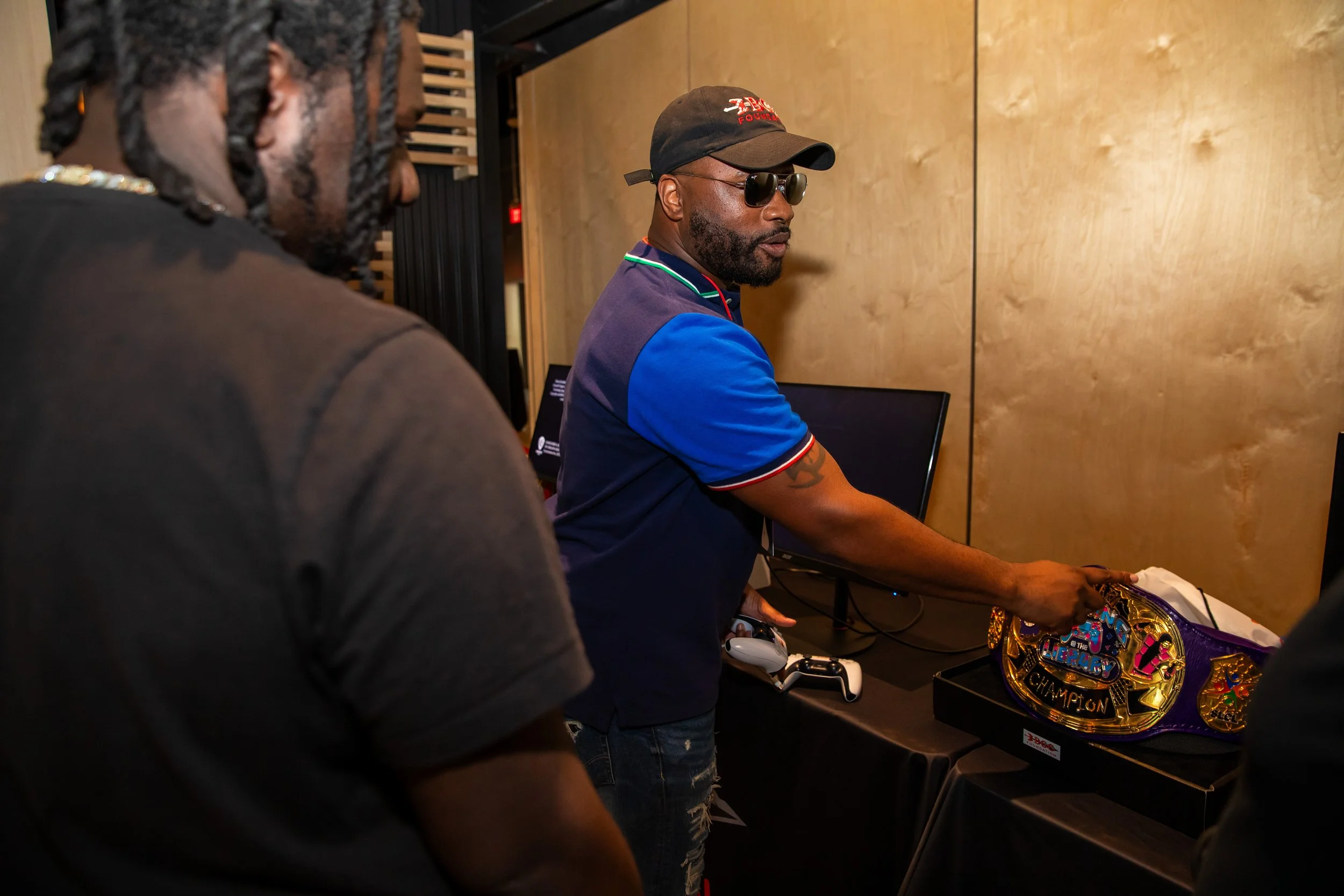 A man wearing sunglasses, a black cap, and a colorful polo shirt is looking at a championship wrestling belt on display. Another person with dreadlocks is partially visible in the foreground. The setting appears to be indoors, possibly at an event or
