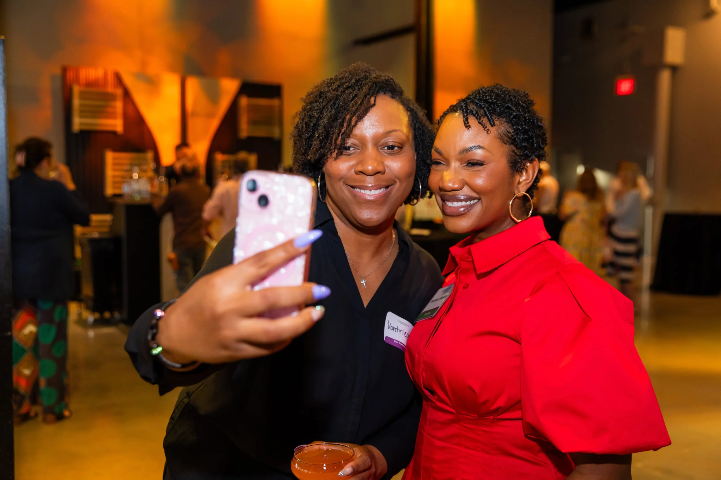 Two women taking a selfie at an indoor event with warm lighting, while others are blurred in the background.