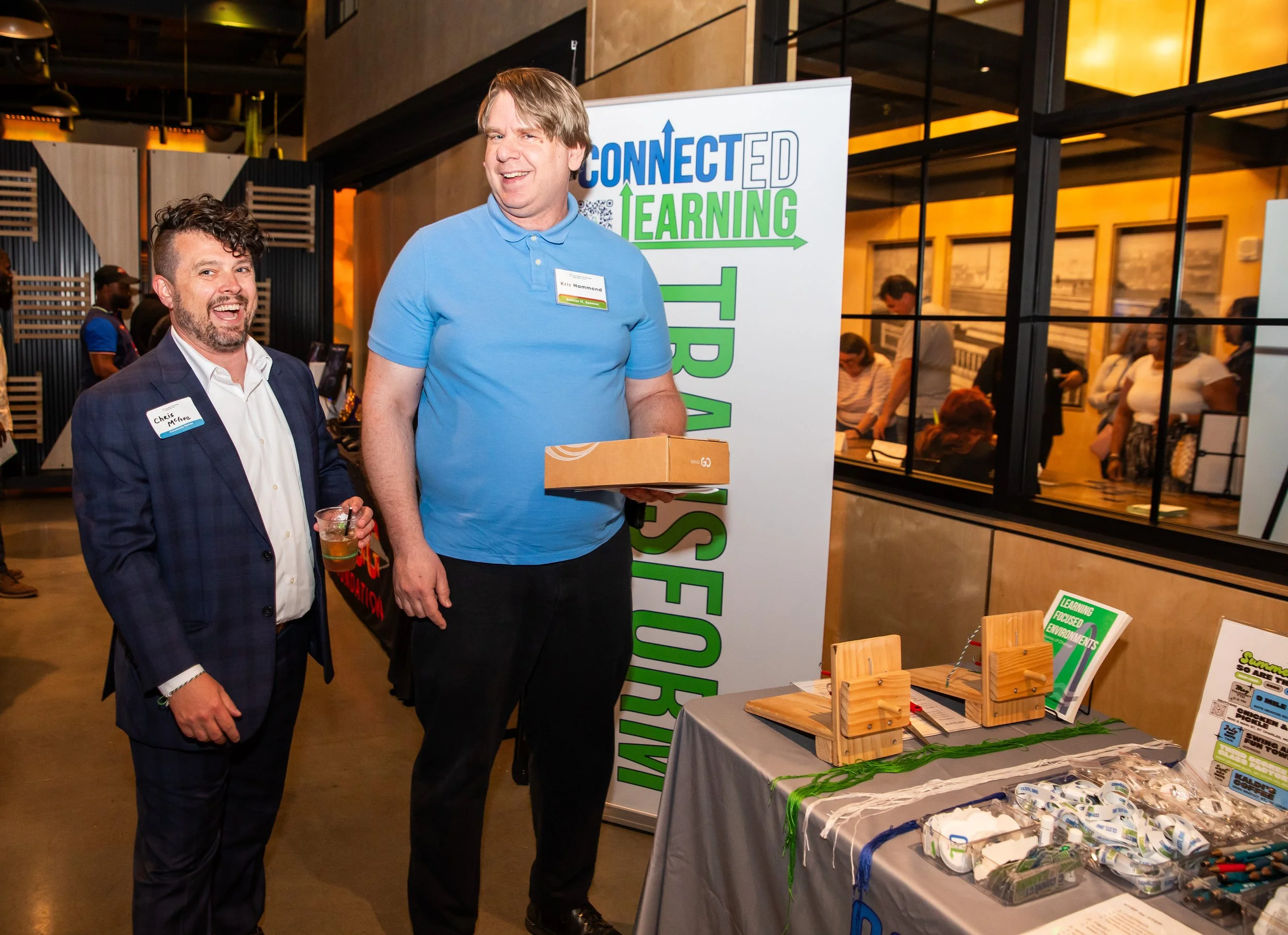 Two men standing at an educational event booth with a sign that reads 'CONNECTED LEARNING'. One man is wearing a suit with a name tag that says 'Chris McAfee' and holding a drink, while the other man is wearing a light blue polo shirt with a name tag