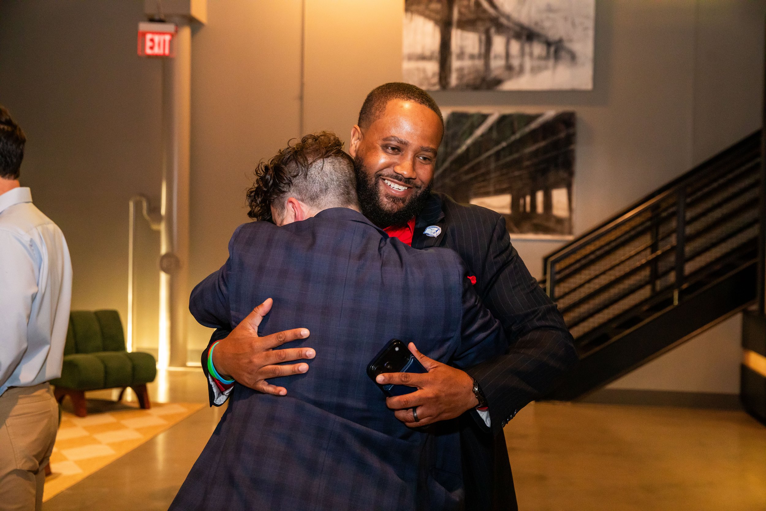 Two men hugging warmly in a modern indoor setting, one holding a smartphone, with artwork and stairs visible in the background.