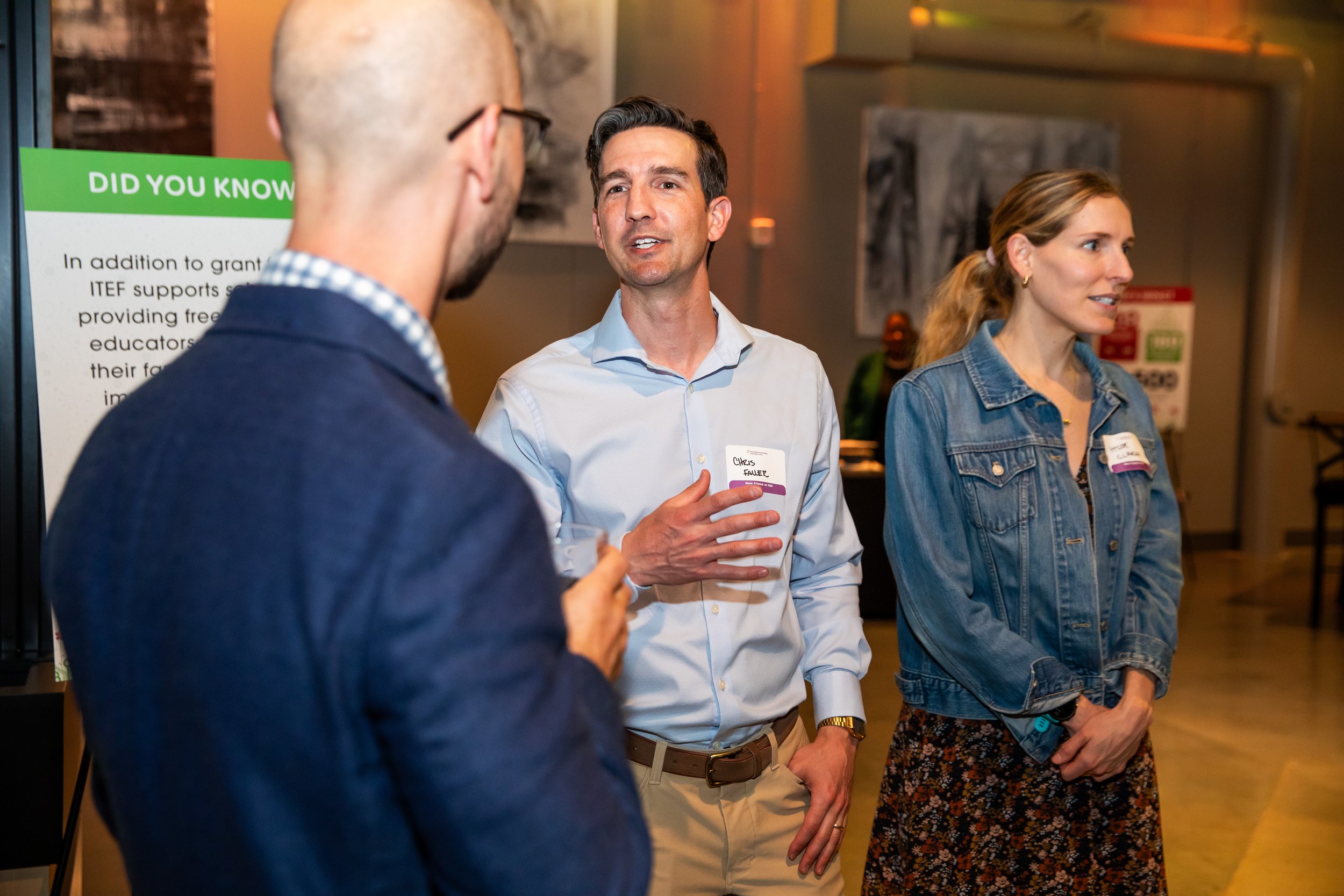 Three people are engaged in conversation at an indoor event. The man in the center is speaking with a name tag that reads 'Chris Faller.' The man on the left is seen from behind, wearing glasses and a navy blazer. The woman on the right has blonde ha