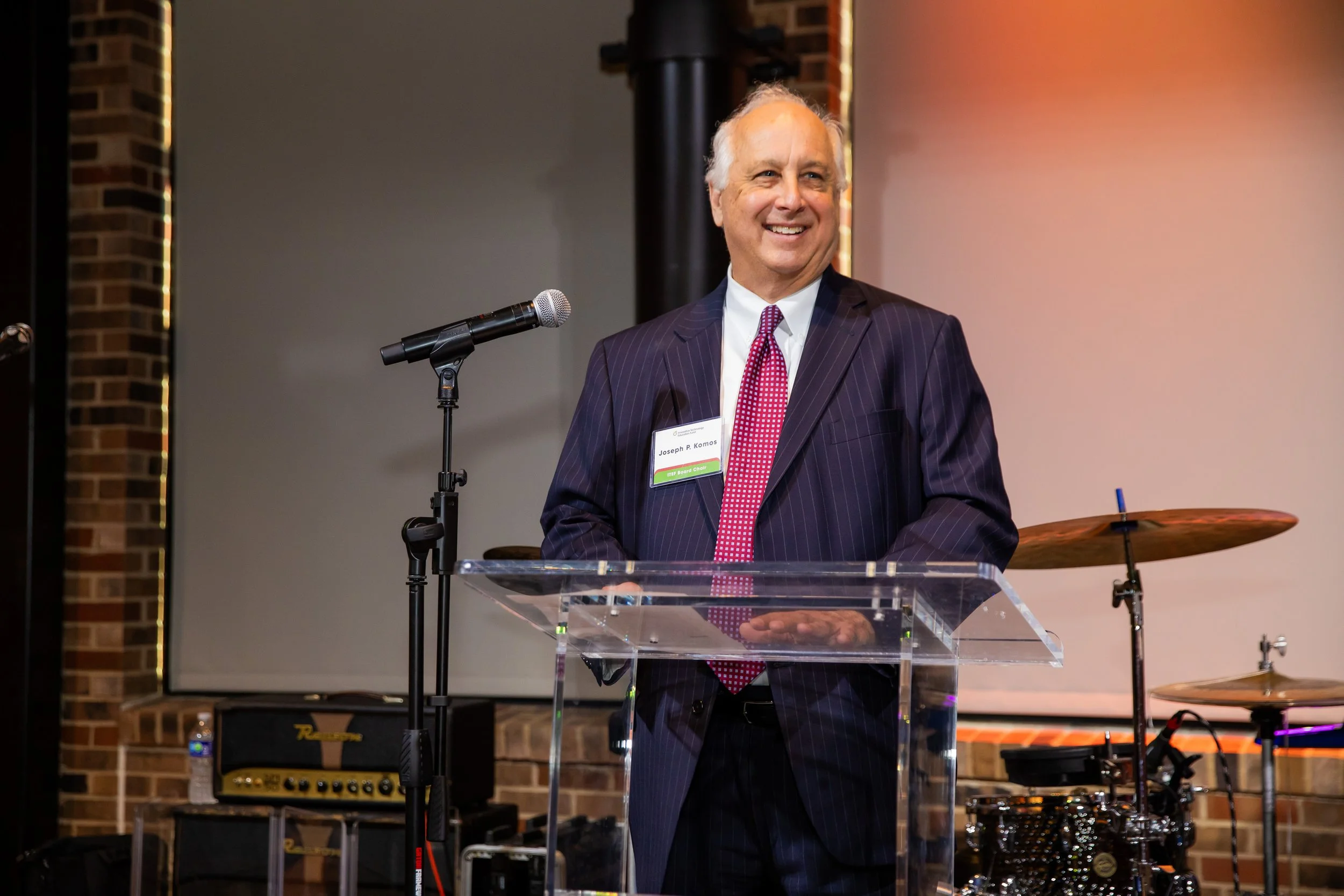 A man in a dark striped suit and red tie is smiling while standing behind a transparent podium with a microphone in a concert or event venue. There is musical equipment, including drums, an amplifier, and a keyboard, in the background.
