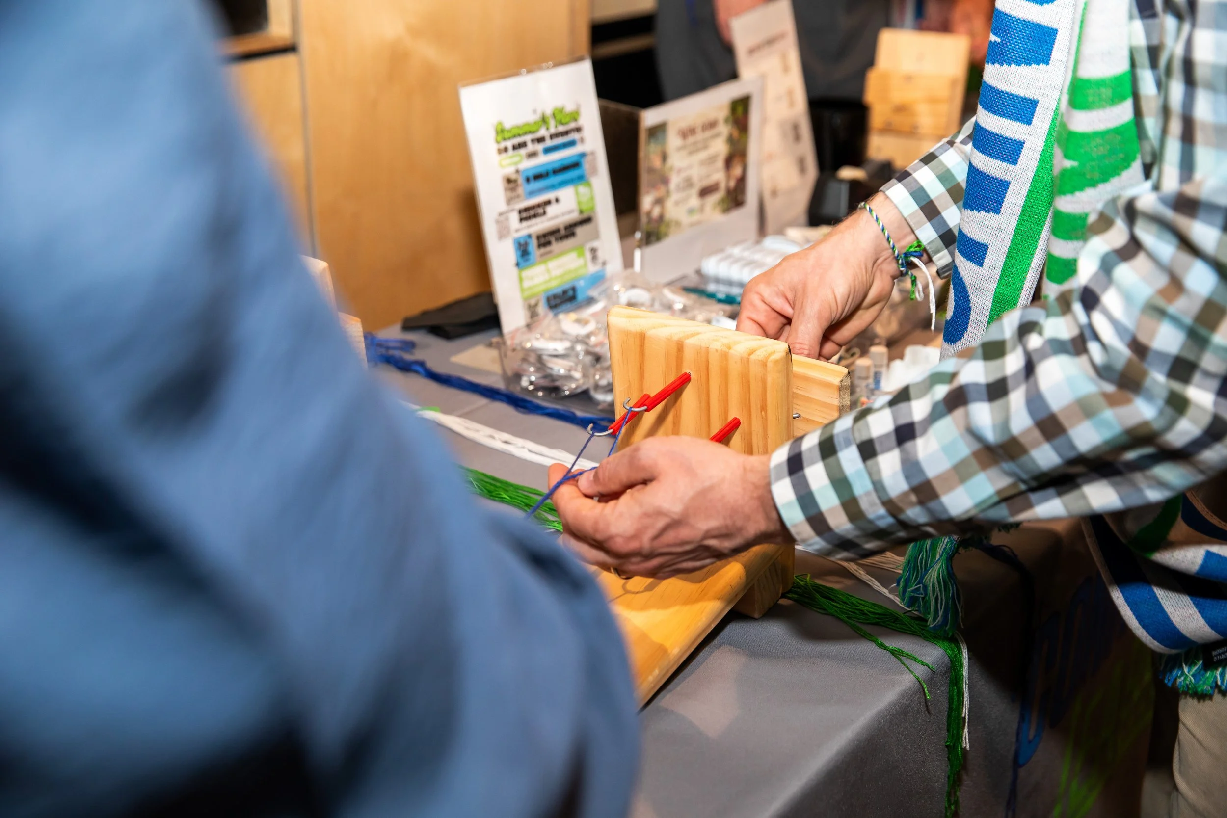People working on a craft project at a table, using colorful yarn and wooden tools at an arts and crafts event.