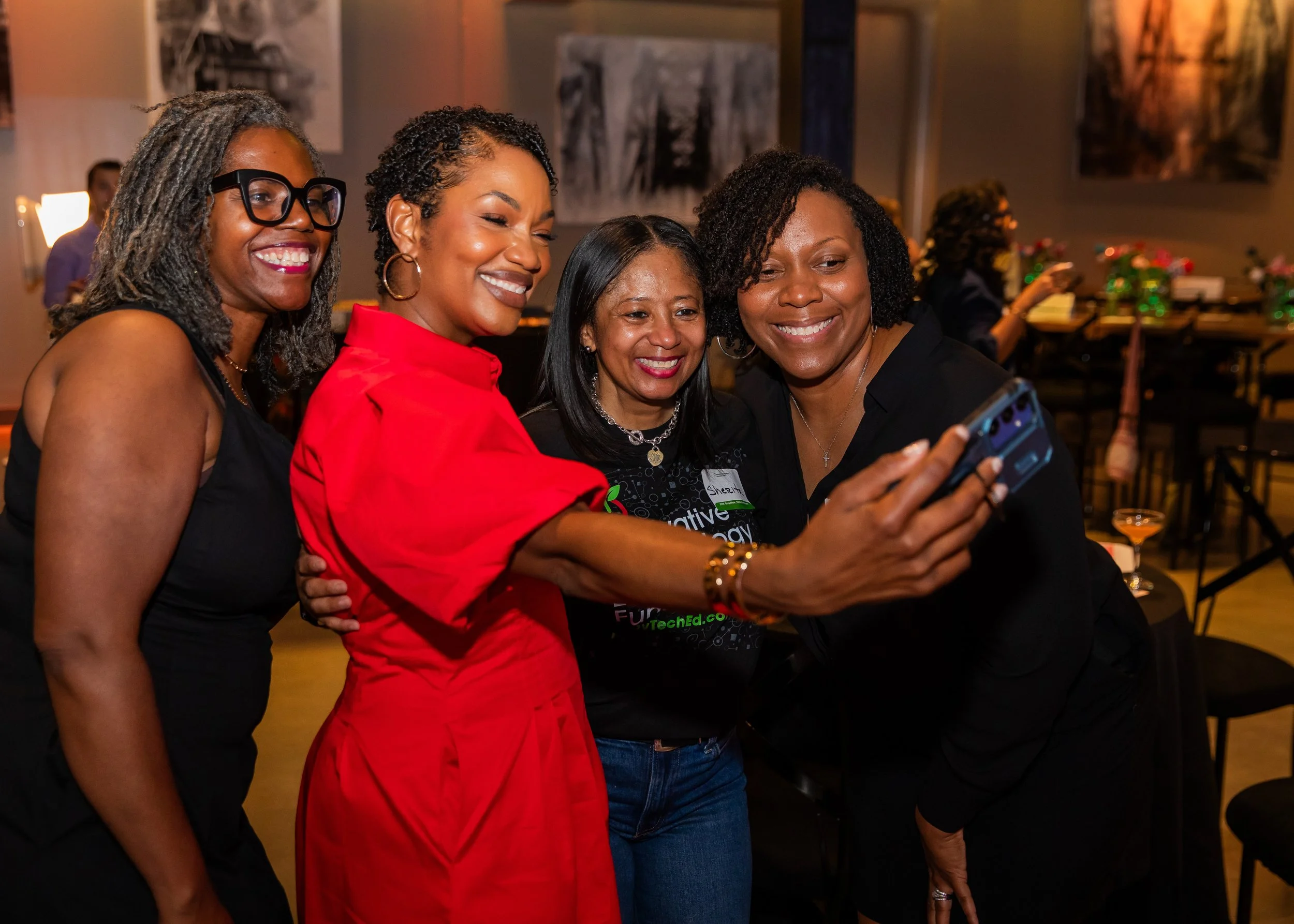 Four women smiling and taking a selfie together at a social gathering or event in an indoor setting.