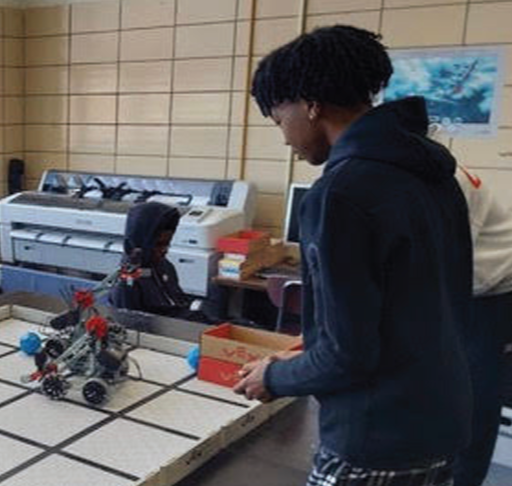 A young man with curly hair in a dark hoodie is looking at a robot on a table in a room with wood-paneled walls. There is a person in a light-colored hoodie in the background near a computer and printer.