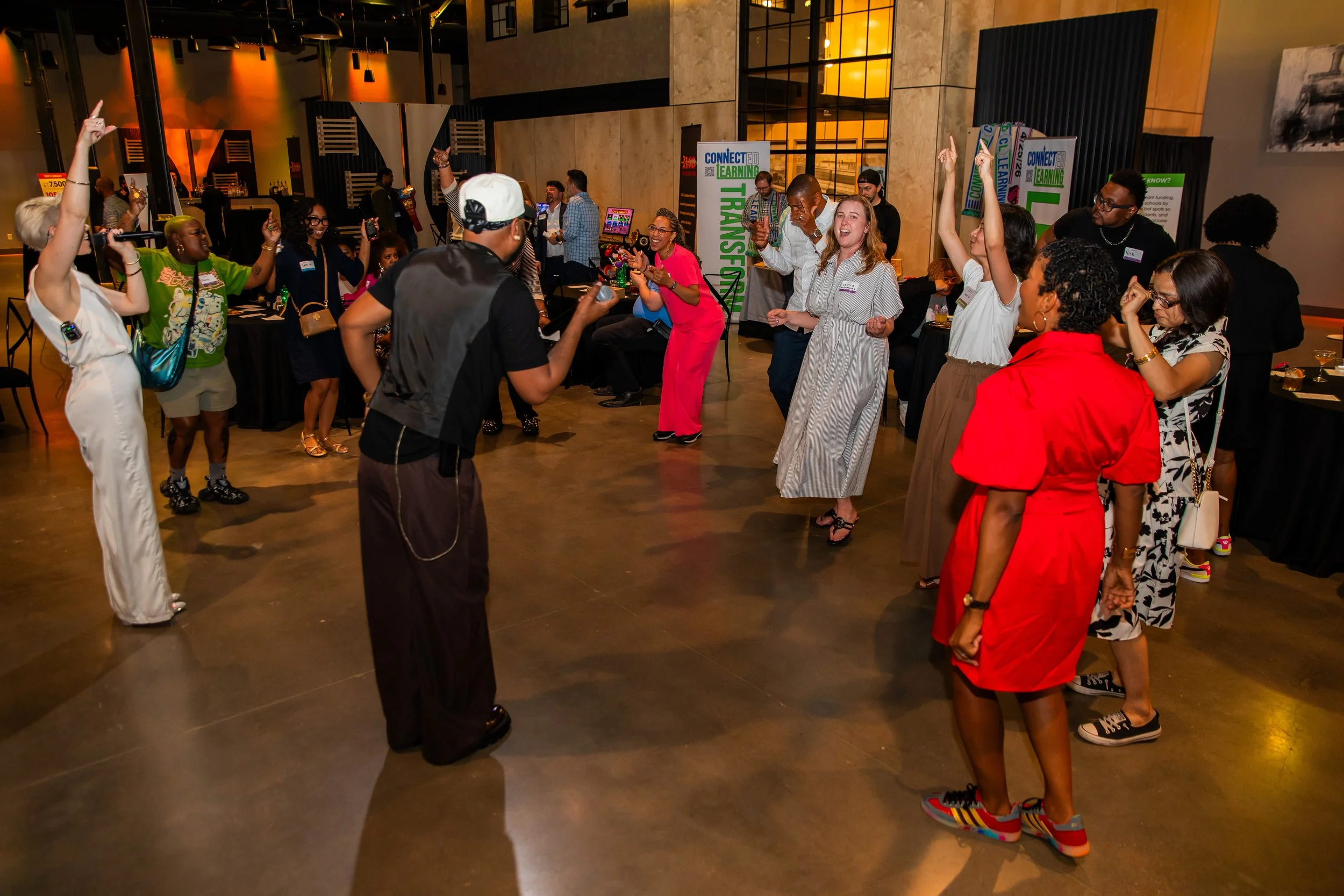 A diverse group of people at an indoor event, engaging in dance and celebration. Some have their hands raised, and others are taking photos or videos. The background shows banners with the words 'CONNECTED LEARNING' and 'TRANSFORMATION,' along with t