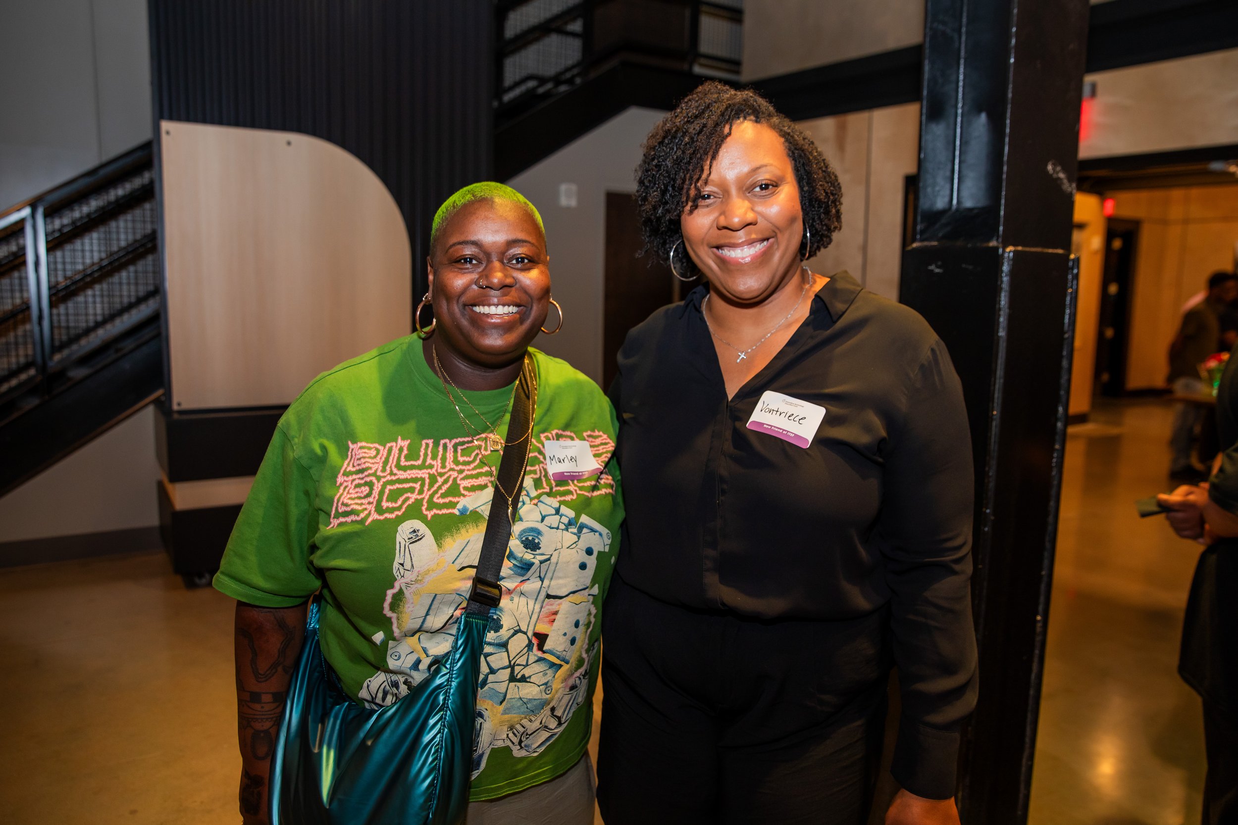 Two women smiling and posing for a photo indoors. One woman has short green hair, wearing a green graphic t-shirt and hoop earrings. The other woman has curly black hair, wearing a black shirt, a necklace with a cross, and a name tag that reads 'Vont