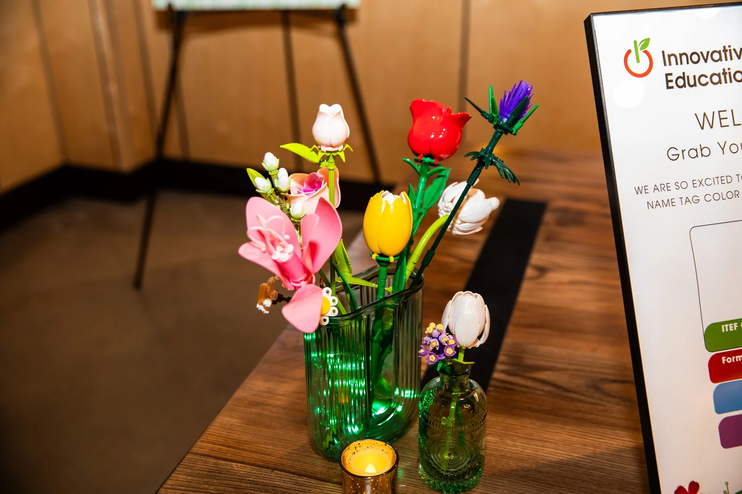 A green glass vase holding various small plastic flowers in colors including pink, white, red, purple, and yellow, placed on a wooden table.