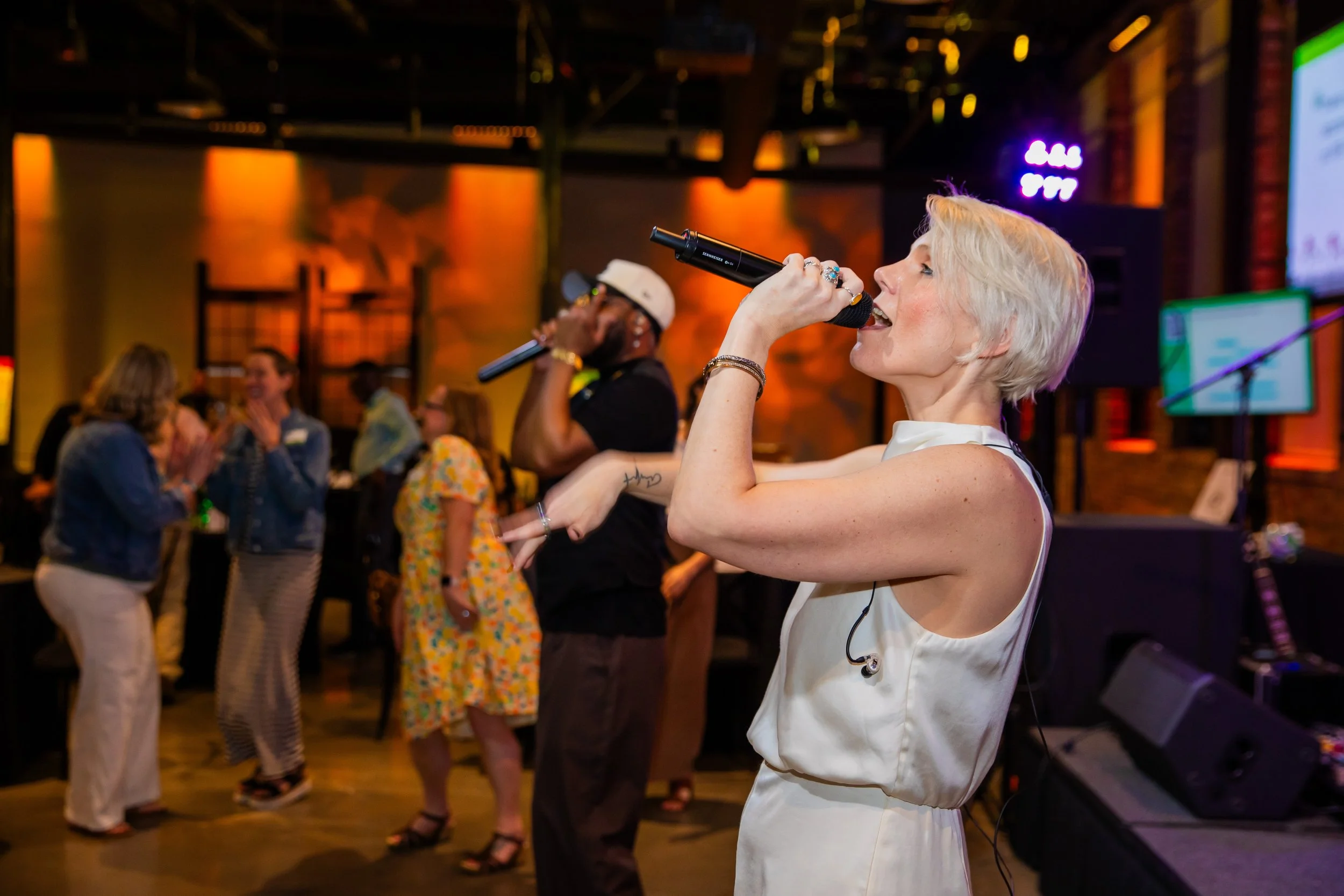 A woman with short blonde hair singing into a microphone at a lively indoor event, with people dancing and socializing in the background.