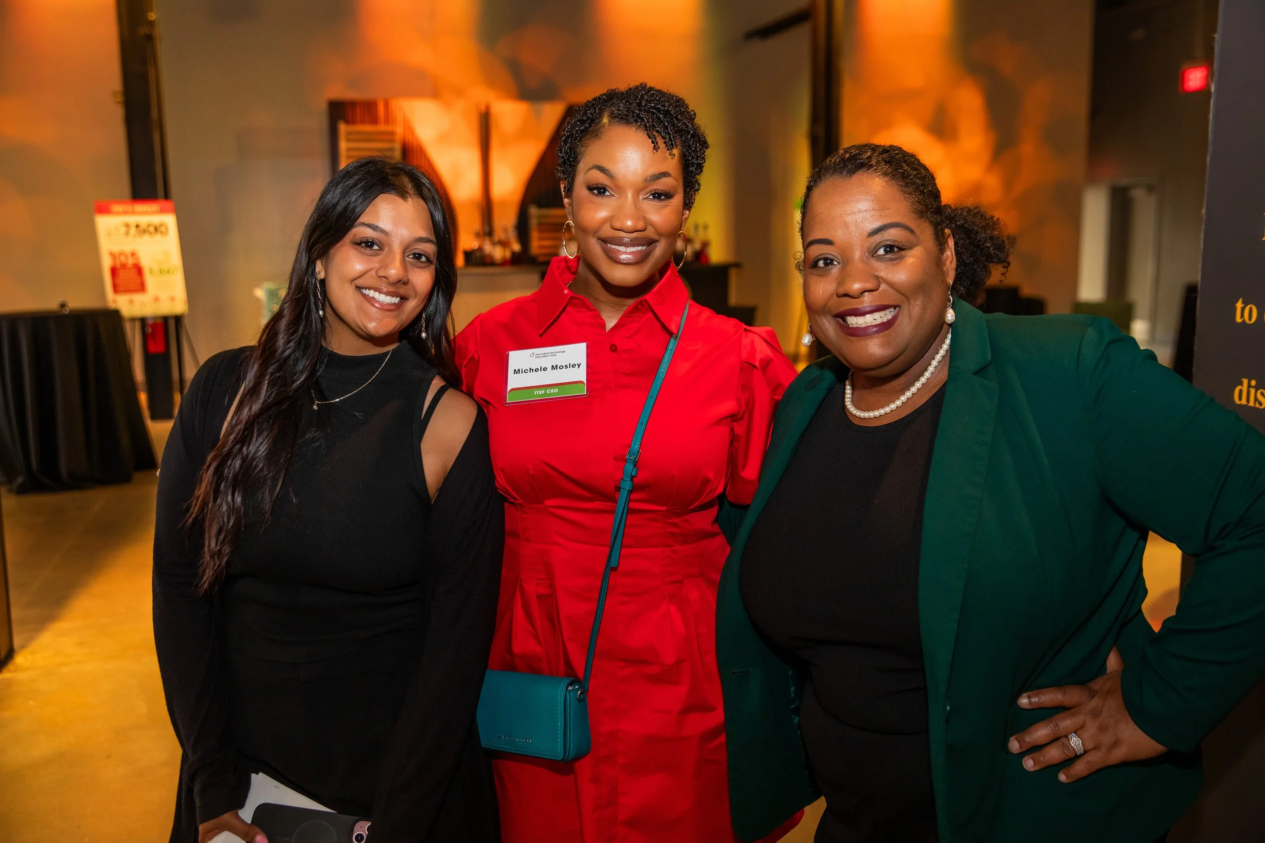 Three women standing together and smiling at a social event or conference. The woman in the middle is wearing a red blouse with a name tag that reads 'Michele Mosley,' and the other two women are wearing black and green attire.