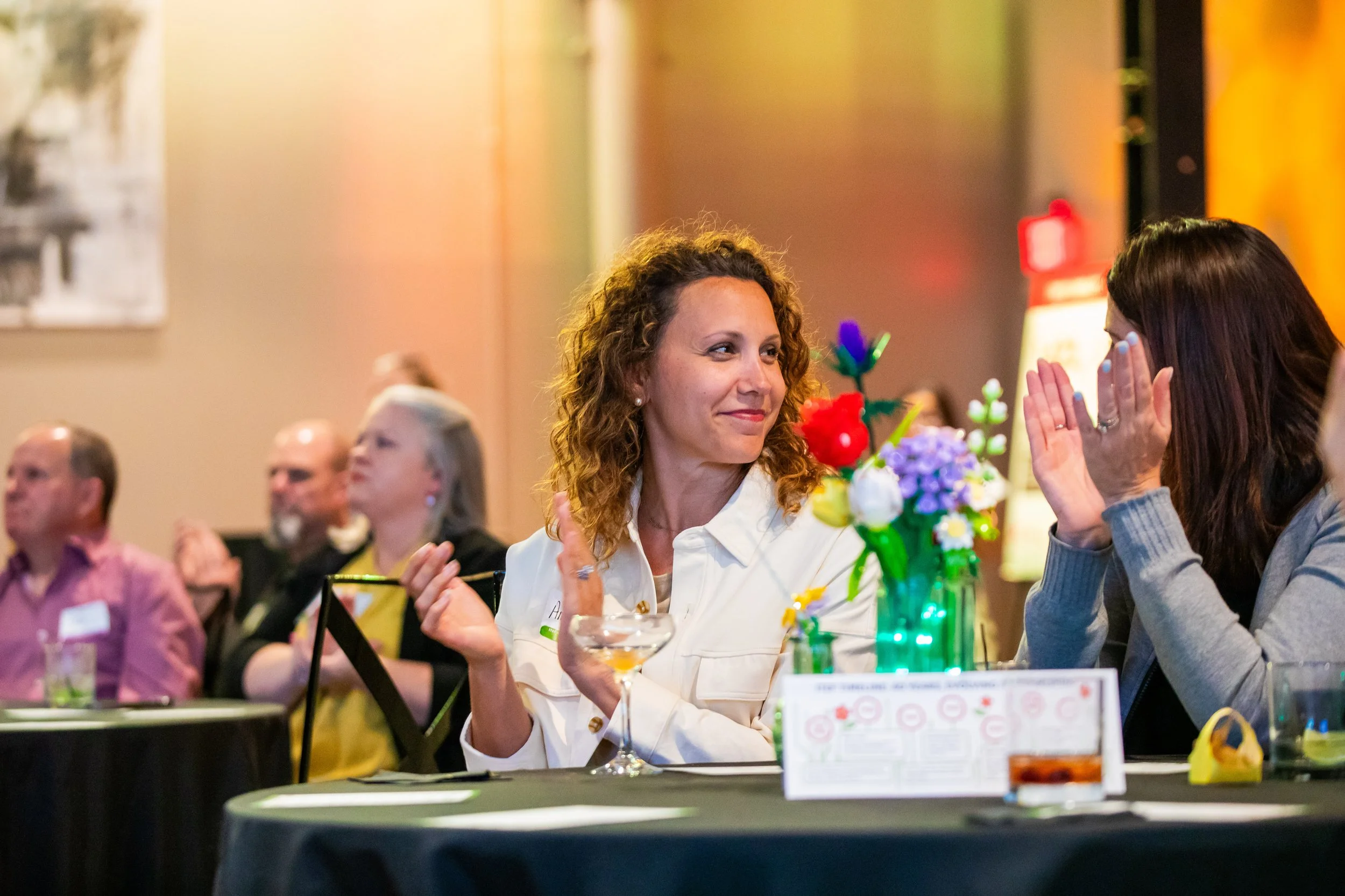 Two women sitting at a table in a conference or banquet setting, engaged in conversation. The woman on the left has curly hair and is wearing a white jacket, smiling. The woman on the right has long dark hair, covering her face with her hands. There 