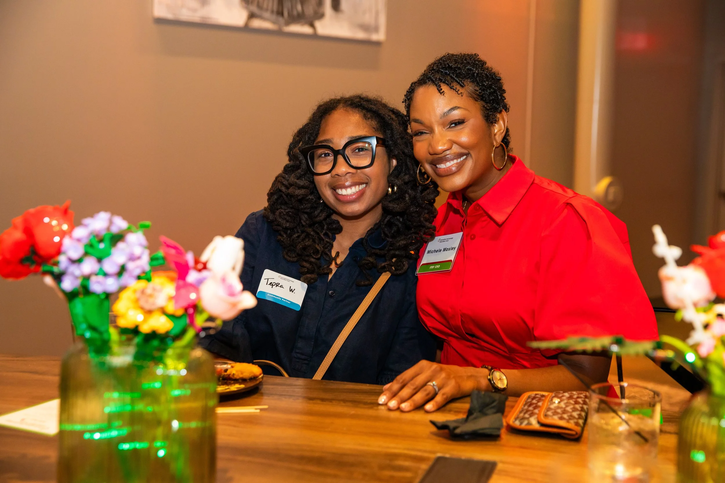 Two women smiling and sitting at a table with colorful flower arrangements in vases. They are wearing name tags and look happy to be together at an indoor event.
