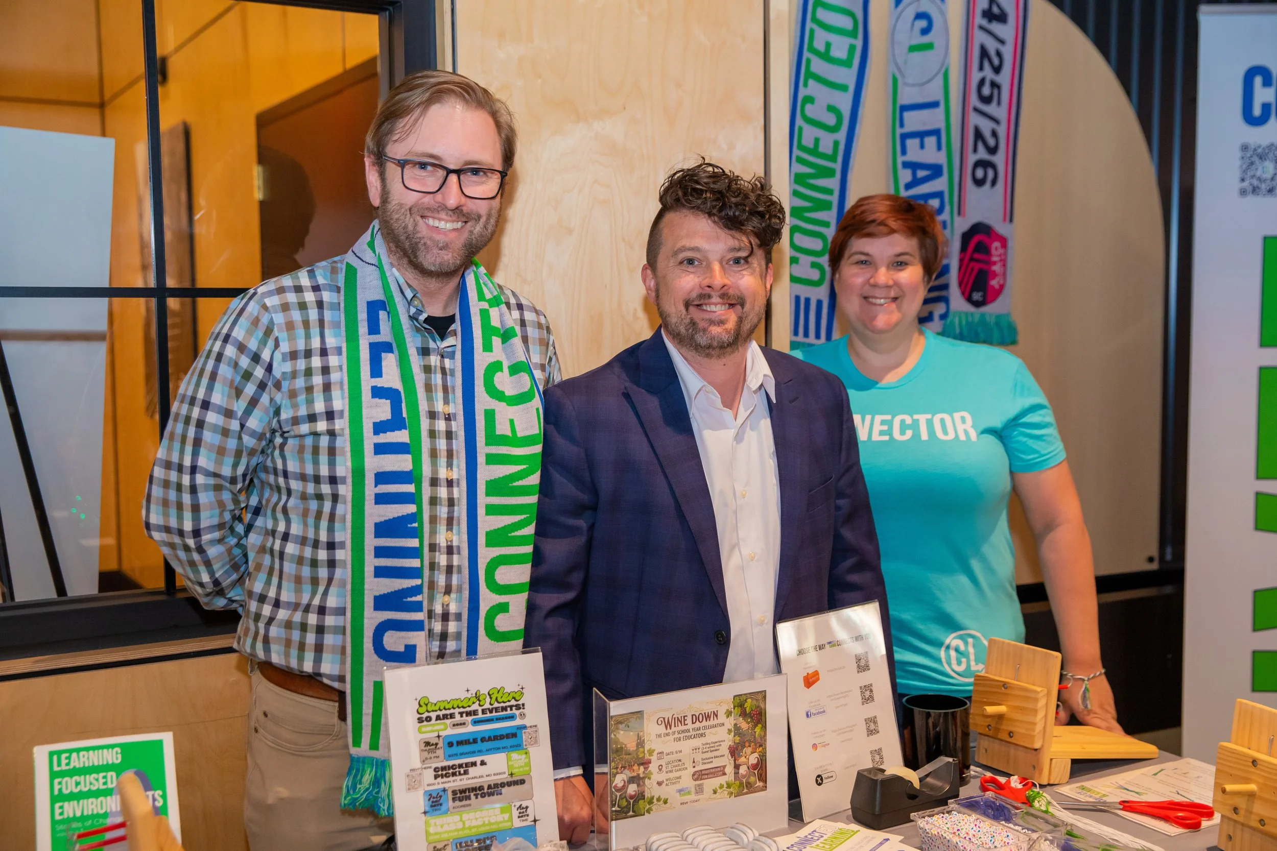 Three people smiling and standing behind a table at an event, with posters and items displayed on the table in front of them.
