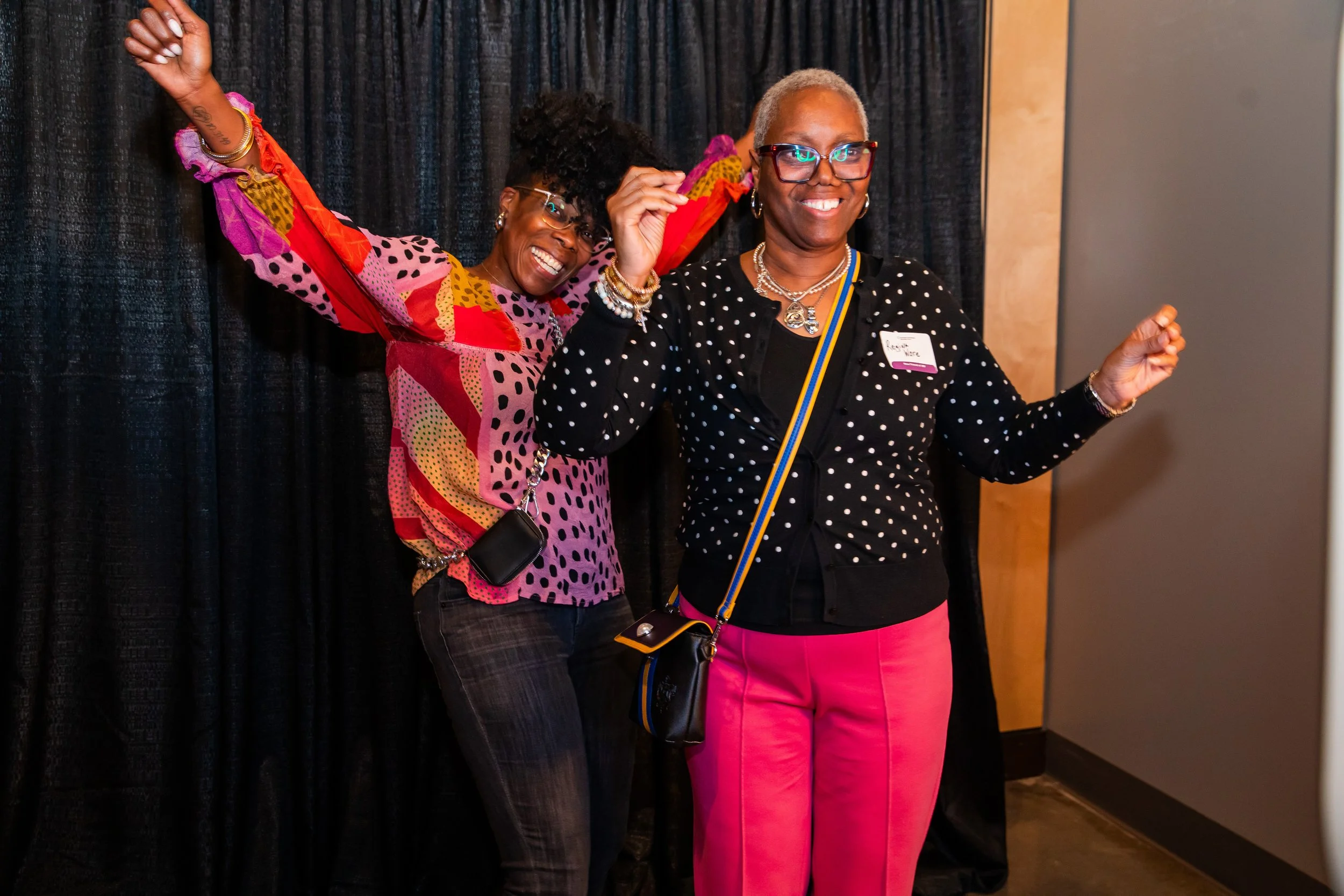 Two women are standing in front of a black curtain, smiling and posing for a photo. The woman on the left has dark skin, glasses, a colorful patchwork top, and dark pants. The woman on the right has lighter skin, short gray hair, glasses, a black pol