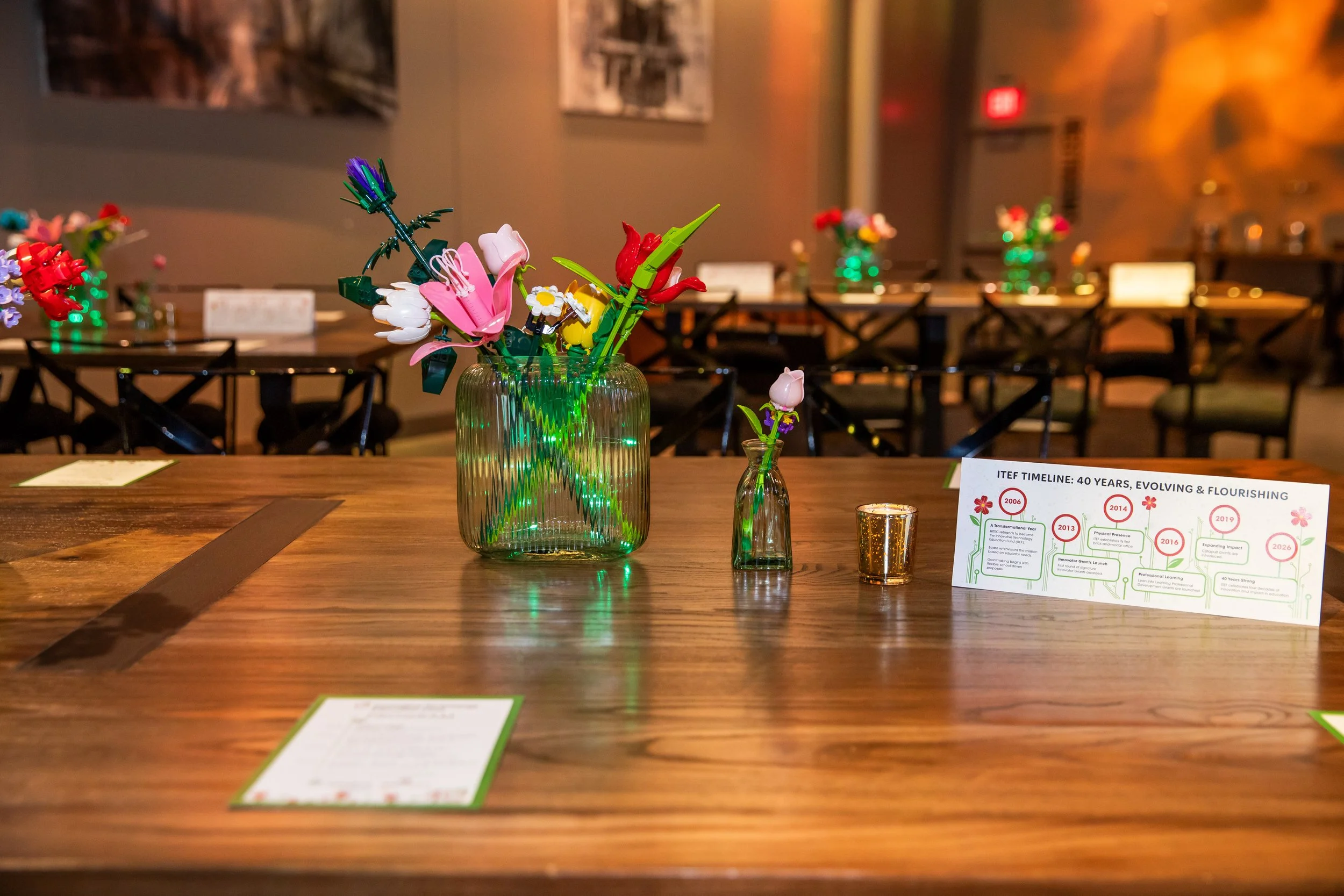 A wooden table with a large glass vase containing colorful artificial flowers, including pink lilies, red tulips, and white daisies, is in a dimly lit room with multiple smaller flower arrangements and informational displays on the table.