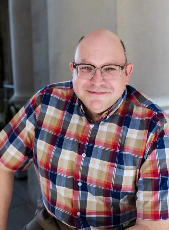 A man with glasses and a friendly smile wearing a colorful plaid shirt, sitting indoors.