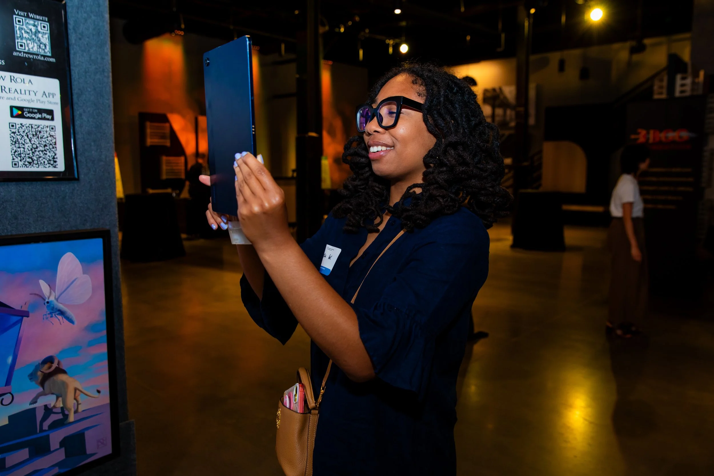 A woman with glasses and curly hair wearing a dark jacket, smiling as she looks at her phone at an indoor event, with digital screens and other people in the background.