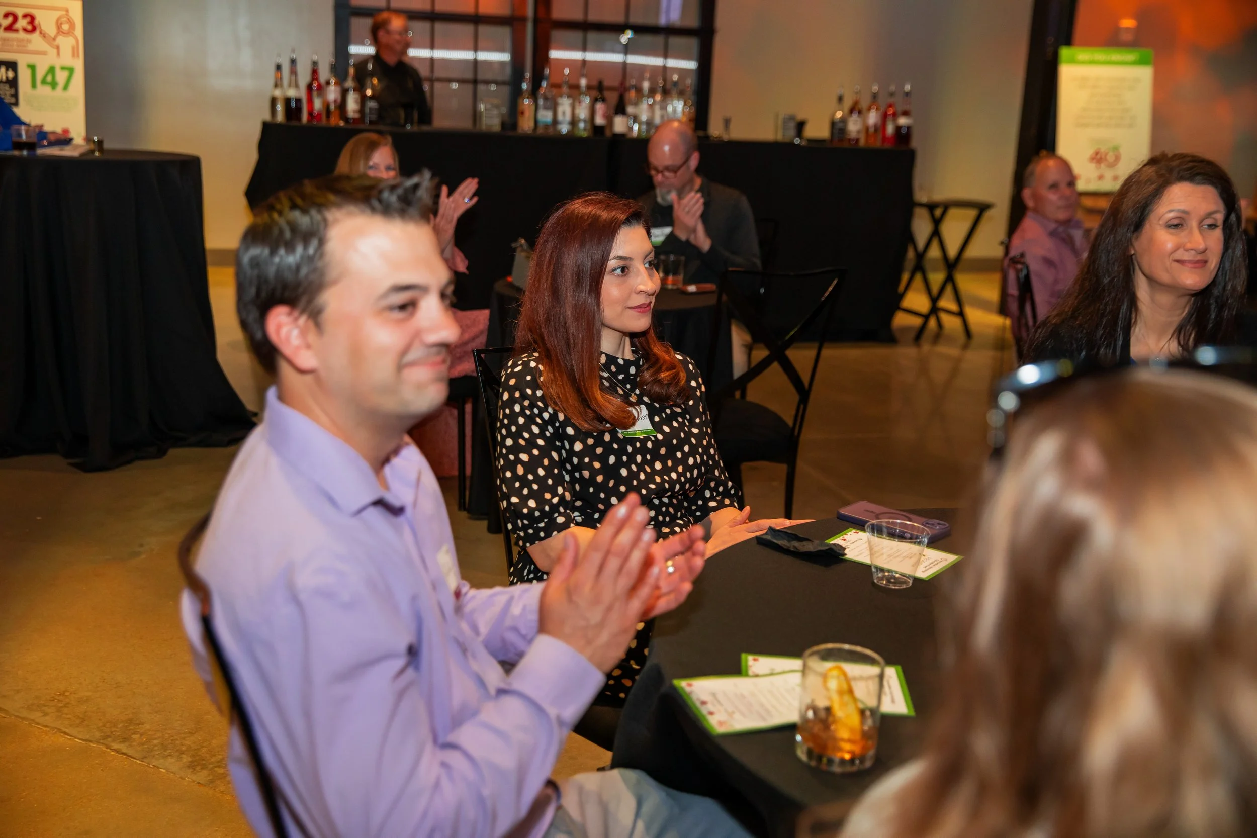 People sitting at tables in a dimly lit venue, with a bar in the background, attending an event or gathering.