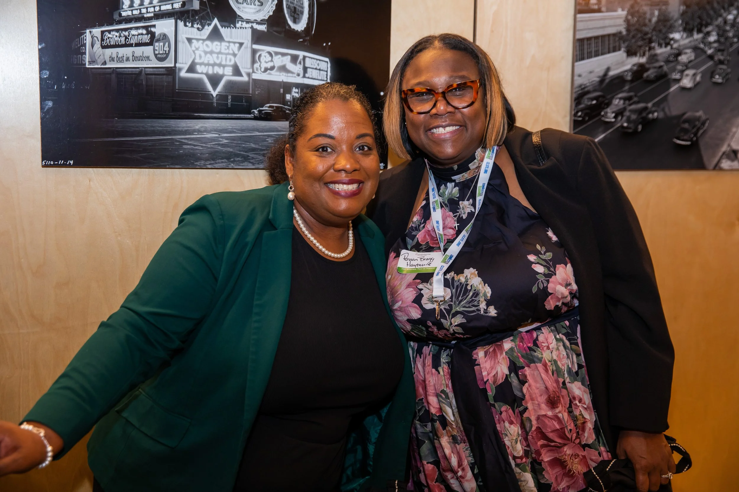 Two women smiling and posing for a photo, standing in front of black and white photographs on a wall. One woman is wearing a green blazer and pearl earrings, while the other woman is wearing glasses and a floral dress with a badge around her neck.
