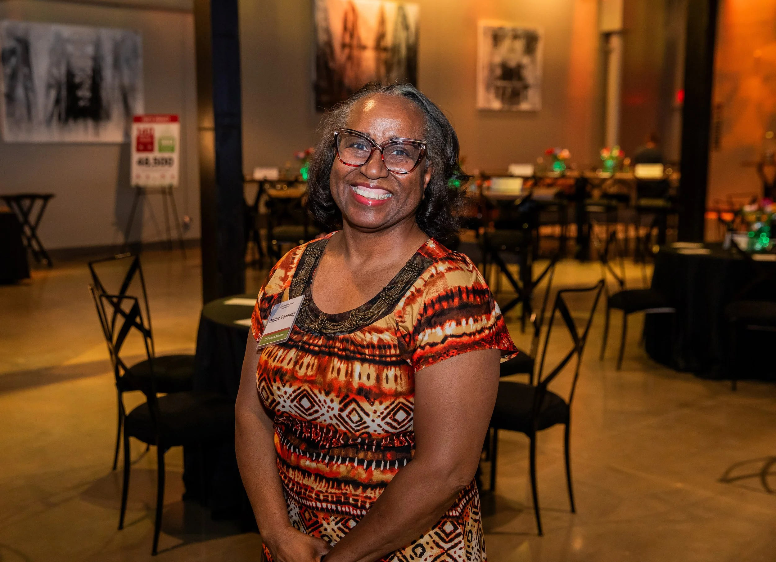 A smiling woman with glasses, wearing a colorful patterned dress, standing in a decorated event space with tables and artwork on the walls.