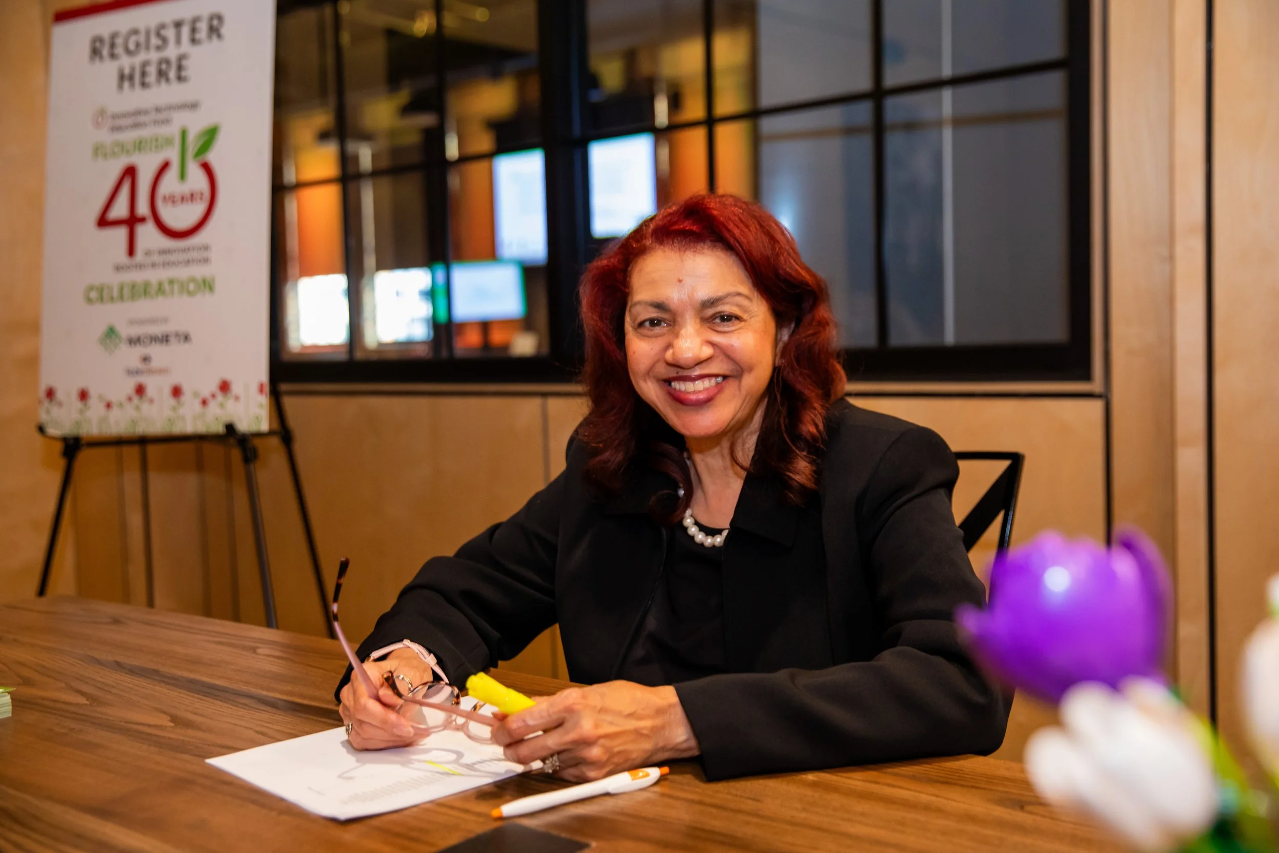 A woman with red hair and a pearl necklace sitting at a wooden table, smiling, with a registration sign in the background.