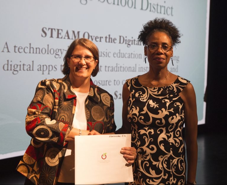 Two women standing together, one holding a certificate, in front of a presentation screen with text about digital education.