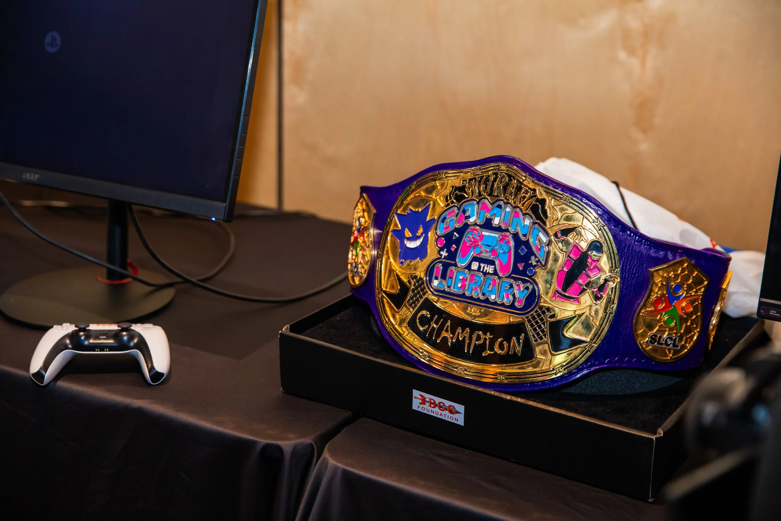 A wrestling championship belt with colorful designs and the words 'Gaming Library Champion' on it, placed on a table next to a gaming controller and two computer monitors.