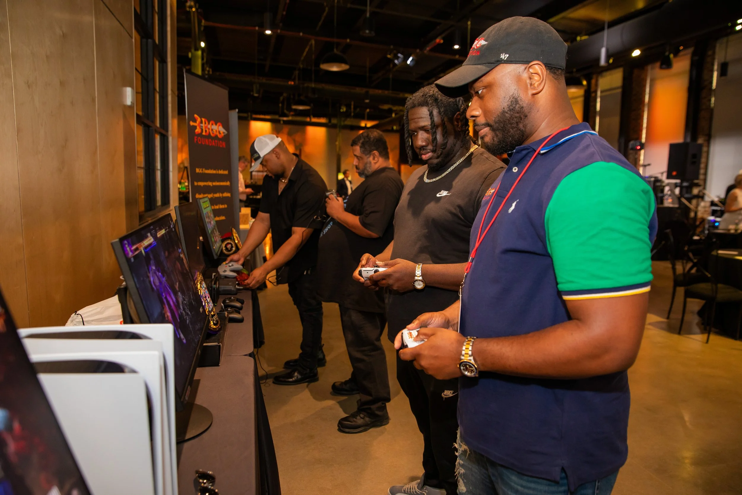 Group of men at a gaming event, playing video games on arcade machines. The setting is indoors with warm lighting and a metal ceiling, and a sign in the background reads 'BGC Foundation'.