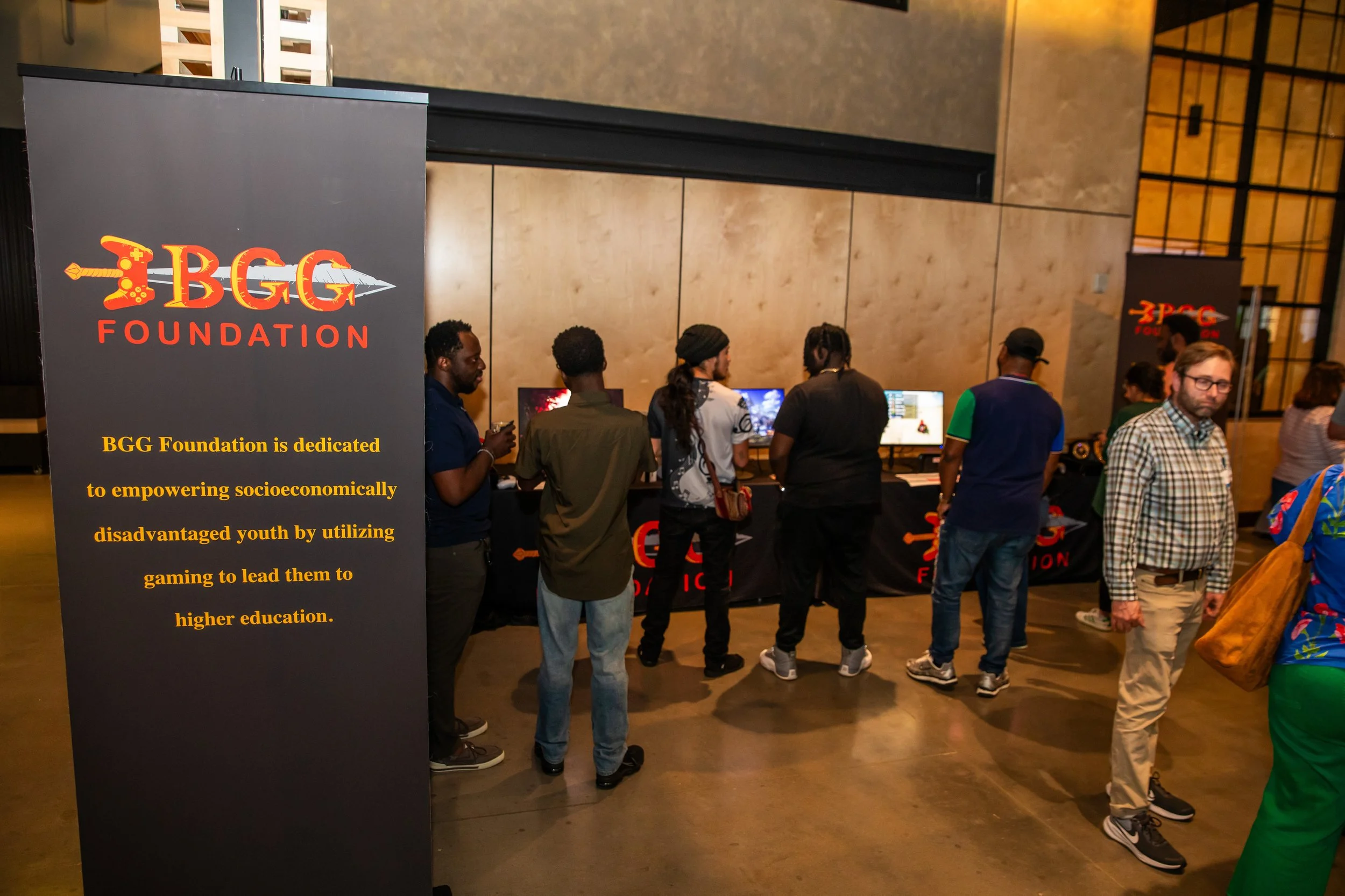 People gathered around gaming stations at a BGG Foundation event with banners and informational displays about empowering socioeconomically disadvantaged youth through gaming.