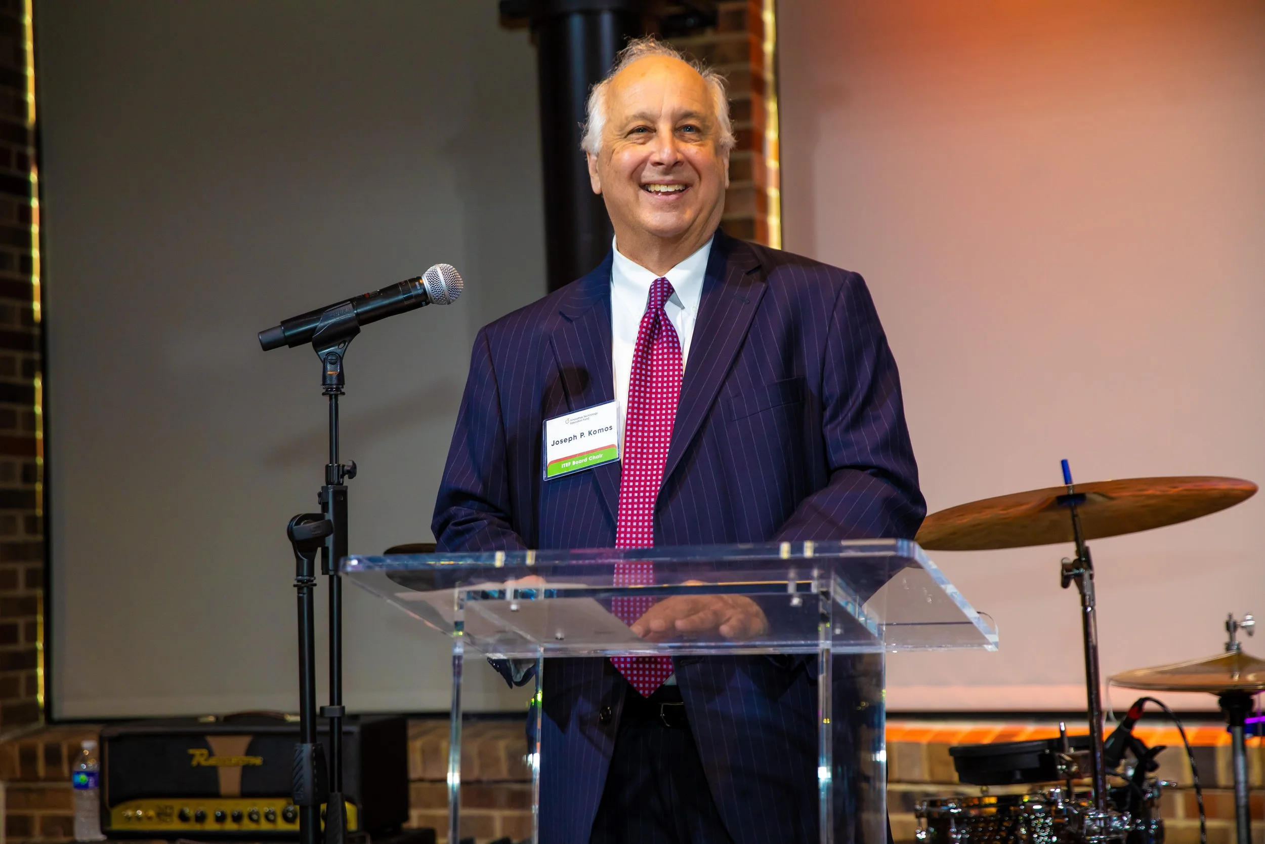 A smiling older man in a dark pinstripe suit and red tie standing behind a clear podium with a microphone in front. He has a name tag that reads Joseph P. Komos and is at a formal event with musical instruments in the background.