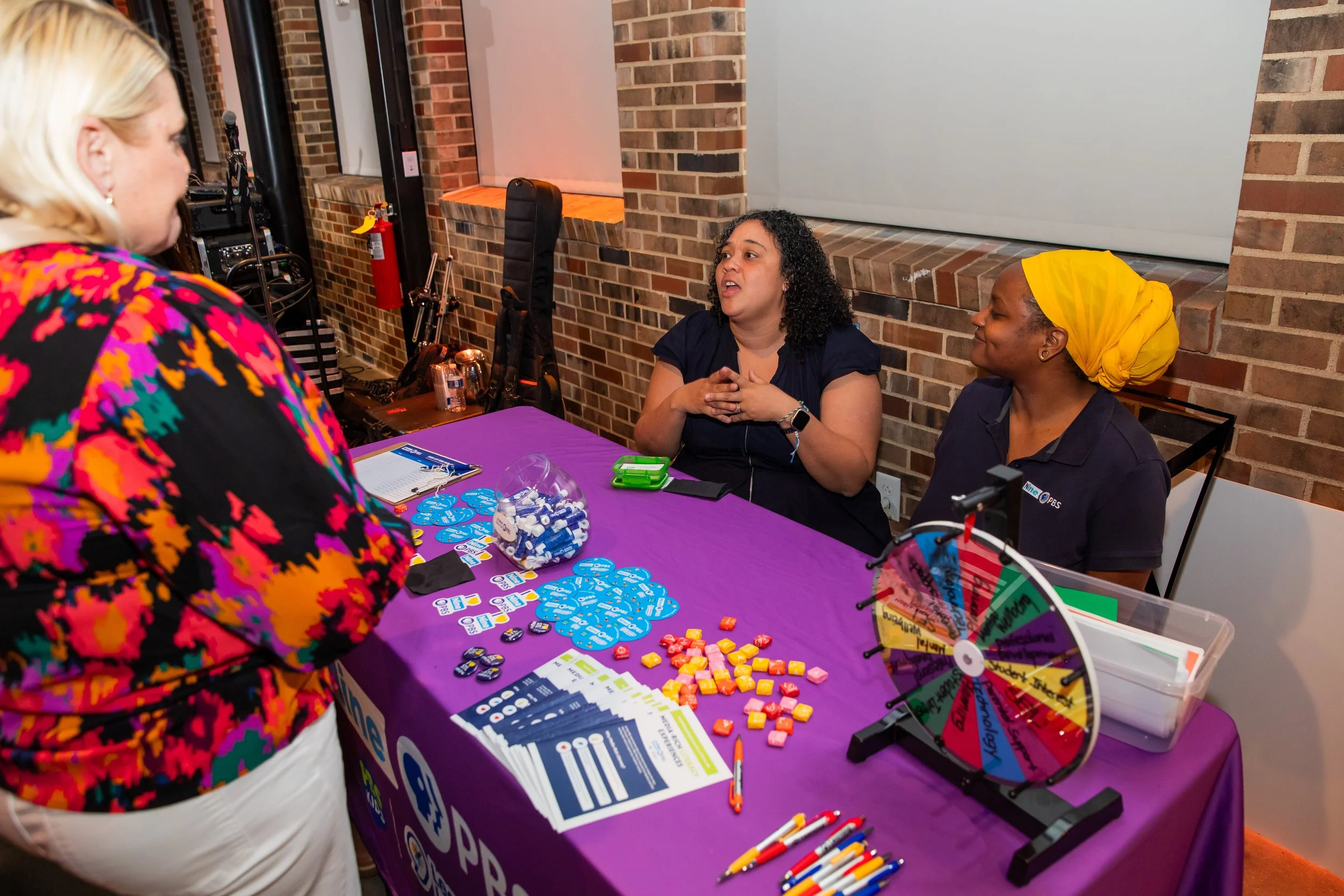 Three women at a booth with a purple tablecloth, discussing a board game or activity involving colorful tokens, cards, and a spinning prize wheel in a brick-walled indoor setting.