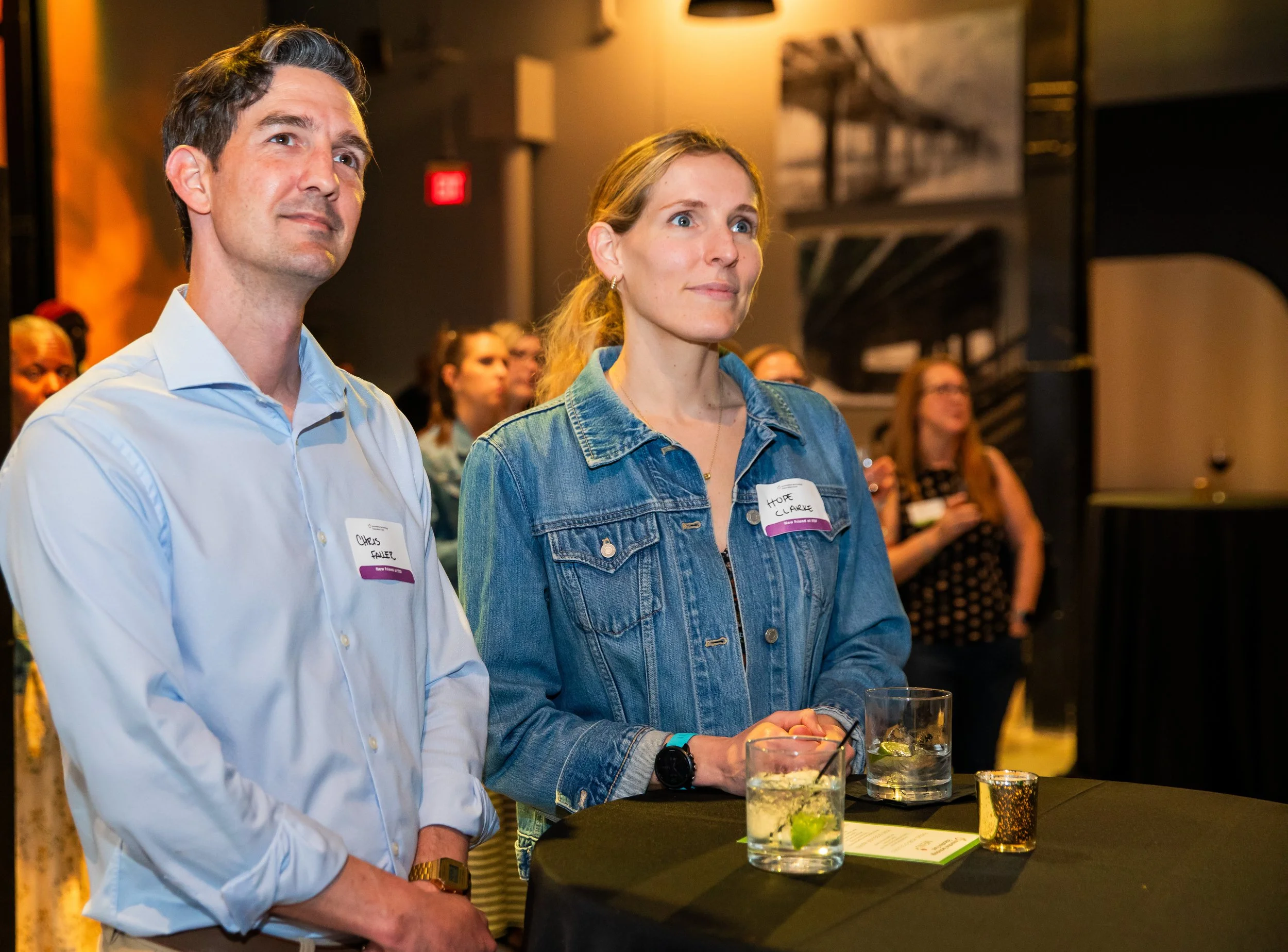 A group of people at a social event, focusing on a man in a white shirt and a woman in a denim jacket, standing near a bar with drinks in front of them.
