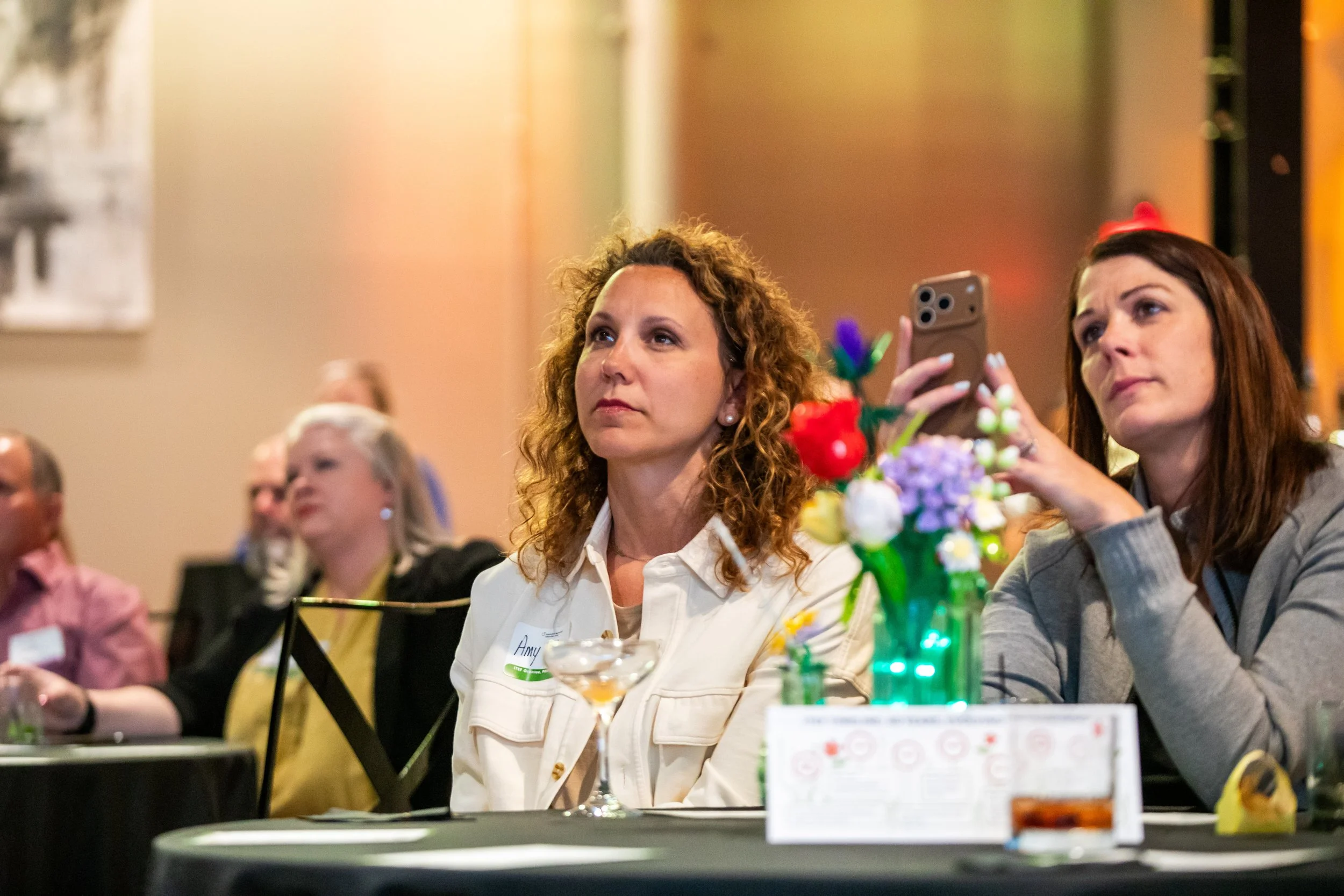 Women attending a formal event or conference, sitting at a table with a colorful floral centerpiece, some attendees are taking photos or listening attentively.
