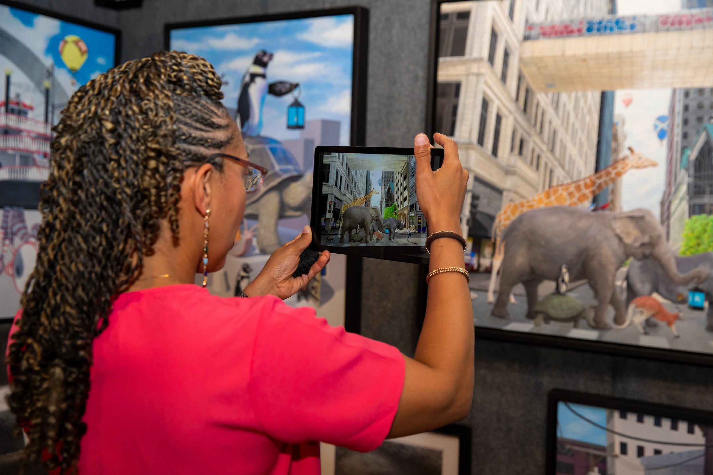 Woman with braided hair wearing a pink shirt takes a photo of a diorama featuring animals including a giraffe, elephants, and a zebra, with city buildings and blue skies in the background.