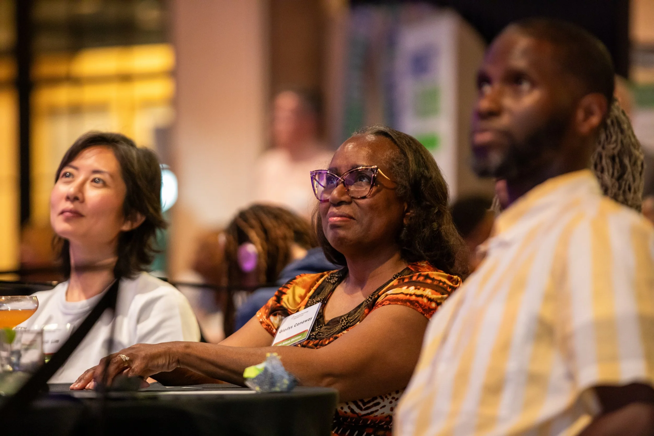 People attentively listening during a conference or seminar, with a diverse audience including Asian, Black, and possibly mixed ethnicity individuals, sitting at tables with drinks and notes.