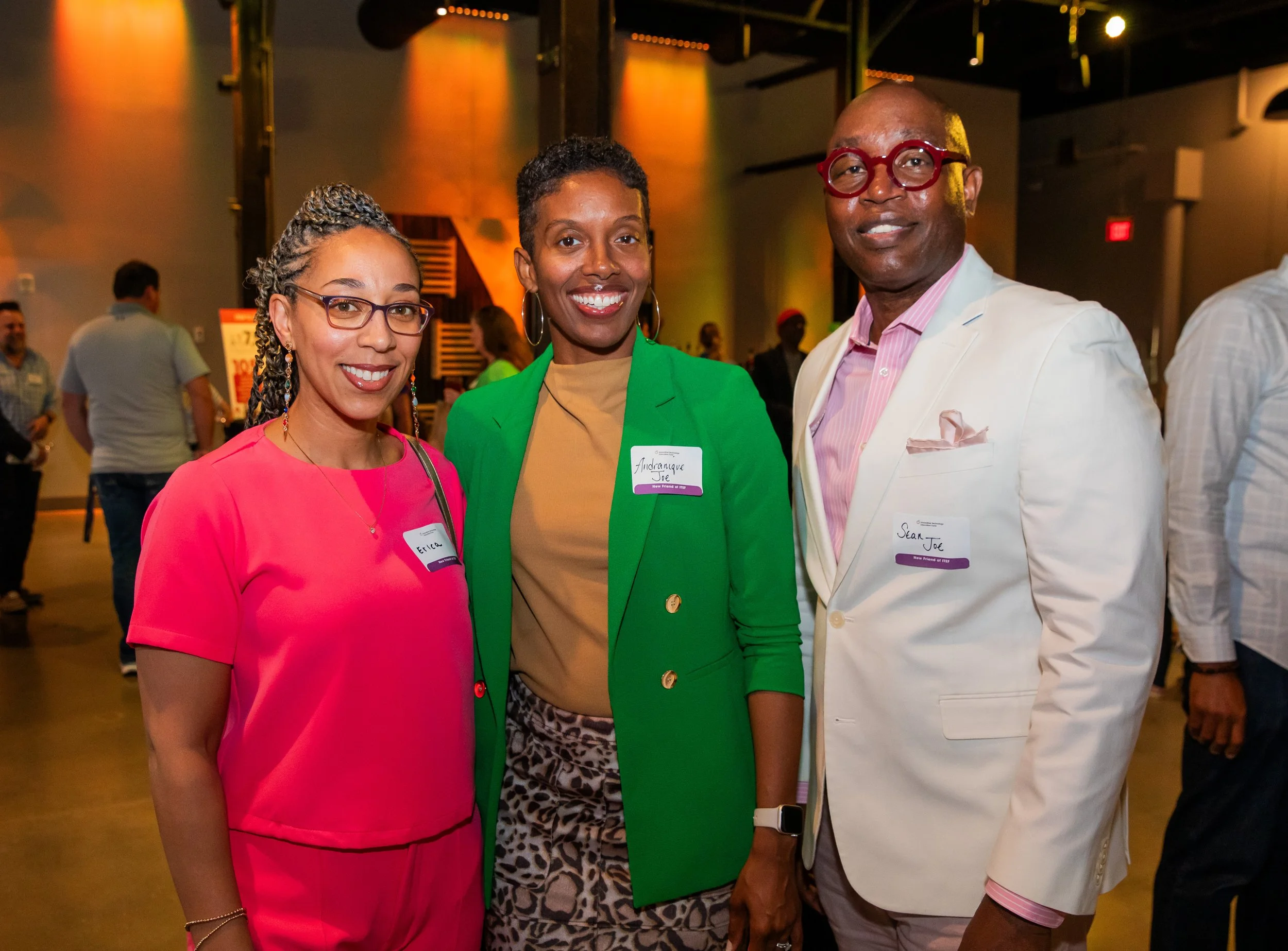 Three people standing together at an indoor event, smiling at the camera. The woman on the left is wearing a bright pink dress and glasses. The woman in the middle is wearing a green blazer and has a cheerful expression. The man on the right is dress