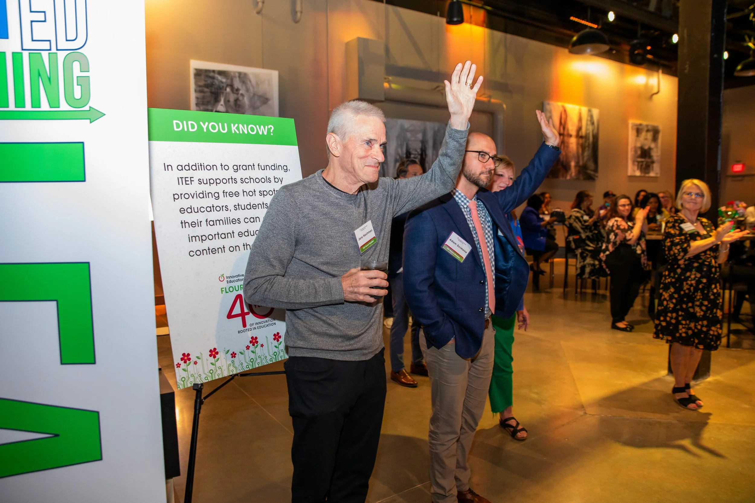 A group of people at an indoor event, with two men in the foreground raising their hands. One man is holding a drink. Behind them, several people are sitting or standing, and a large colorful sign with a green and white design displays informational 