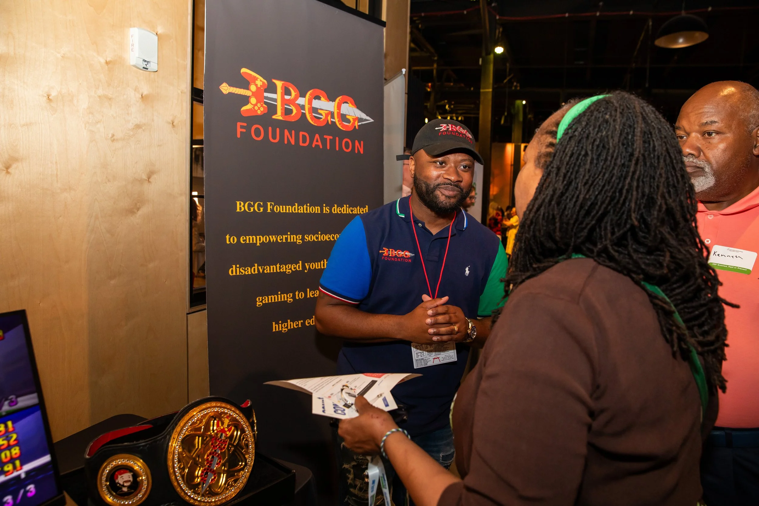 A man wearing a cap and a polo shirt with a logo is speaking to two women at a booth of the BGG Foundation. The booth display includes a banner with the foundation's logo and information, as well as some gaming trophies and a brochure on the table.